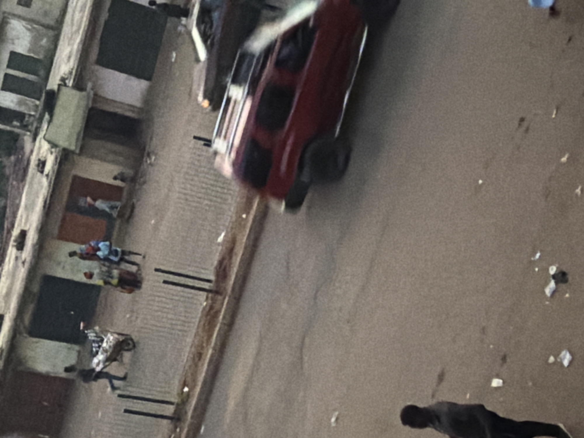 In Bukuru, Nigeria, a street scene unfolds with several individuals and vehicles. In the foreground, a dark-skinned man stands facing away from the camera, his stance suggesting he is observing the surroundings. Further down the street, a red SUV with a roof rack is in motion, its blurred appearance indicating movement. To the left of the SUV, a darker colored sedan is also visible, partially obscured by the SUV. On the sidewalk or road adjacent to the buildings, a person is pushing a wheelbarrow laden with goods, accompanied by two other individuals, one of whom appears to be carrying a child on their back. The buildings in the background have shuttered windows and doors, suggesting a commercial or residential area. Scattered litter is present on the ground, typical of an urban environment.