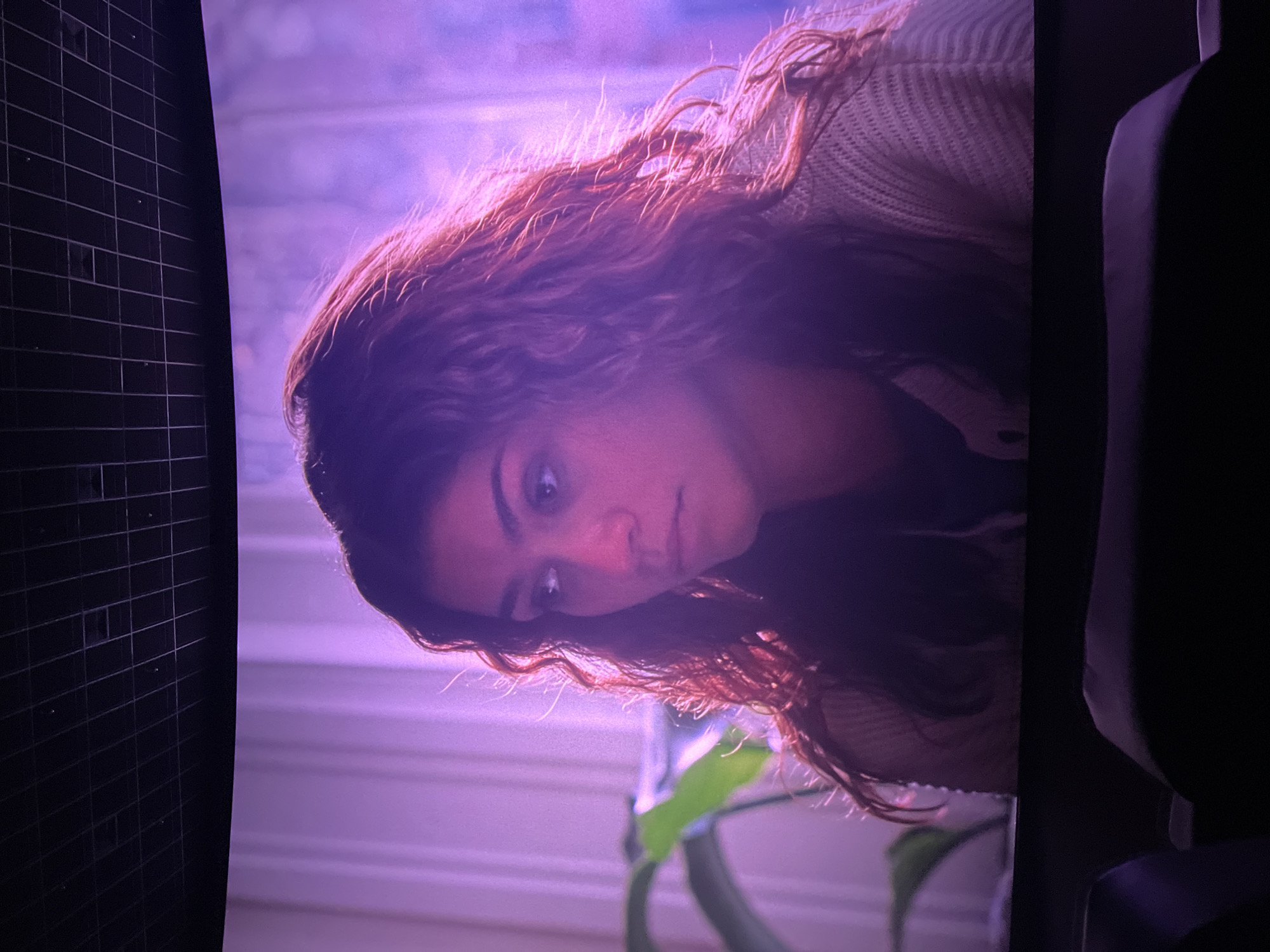 The image features a young woman looking downwards, captured from the chest up. She has long, wavy reddish-brown hair, illuminated by a strong purple-pink light, especially around her head and shoulders. Her eyes are wide, gazing towards the lower left of the frame (when oriented correctly), with an expression that suggests curiosity, concern, or contemplation. She is wearing a light-colored, possibly cream or beige, knitted top.
The setting appears to be indoors. The background is dominated by a soft purple-pink glow, possibly from a screen or artificial lighting, and shows faint horizontal lines that could indicate blinds or a textured wall. A portion of a green-leafed plant is visible in the lower left background. On the far left edge of the image, a dark, grid-like pattern is present, resembling a screen or a window covering. The far right edge shows a dark, indistinct shape, possibly part of a chair or furniture. The lighting is generally dim, with the purple hue creating a distinct atmosphere. No text is visible in the image. While a location of Khimki, Russia was provided, there are no visual cues within the image itself to confirm this.