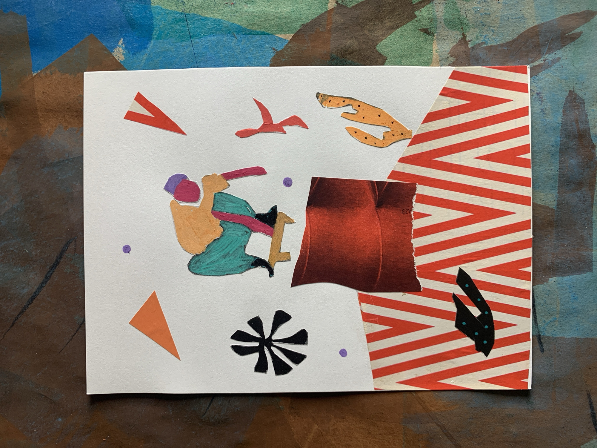 This is a collage artwork created on white paper, depicting a stylized figure in mid-air on a skateboard. The figure is wearing orange and teal clothing and a purple head covering. Around the figure are various abstract shapes and elements, including orange triangles, red shapes, a black asterisk-like symbol, purple dots, and an orange, cactus-like shape. The lower portion of the collage features a section of red and white striped paper. The artwork is placed on a textured surface with a mix of brown, blue, and green colors, suggesting it is a background or base for the collage. The image is a still life of the collage itself, with no indication of a specific location in Sergeevo, Russia.