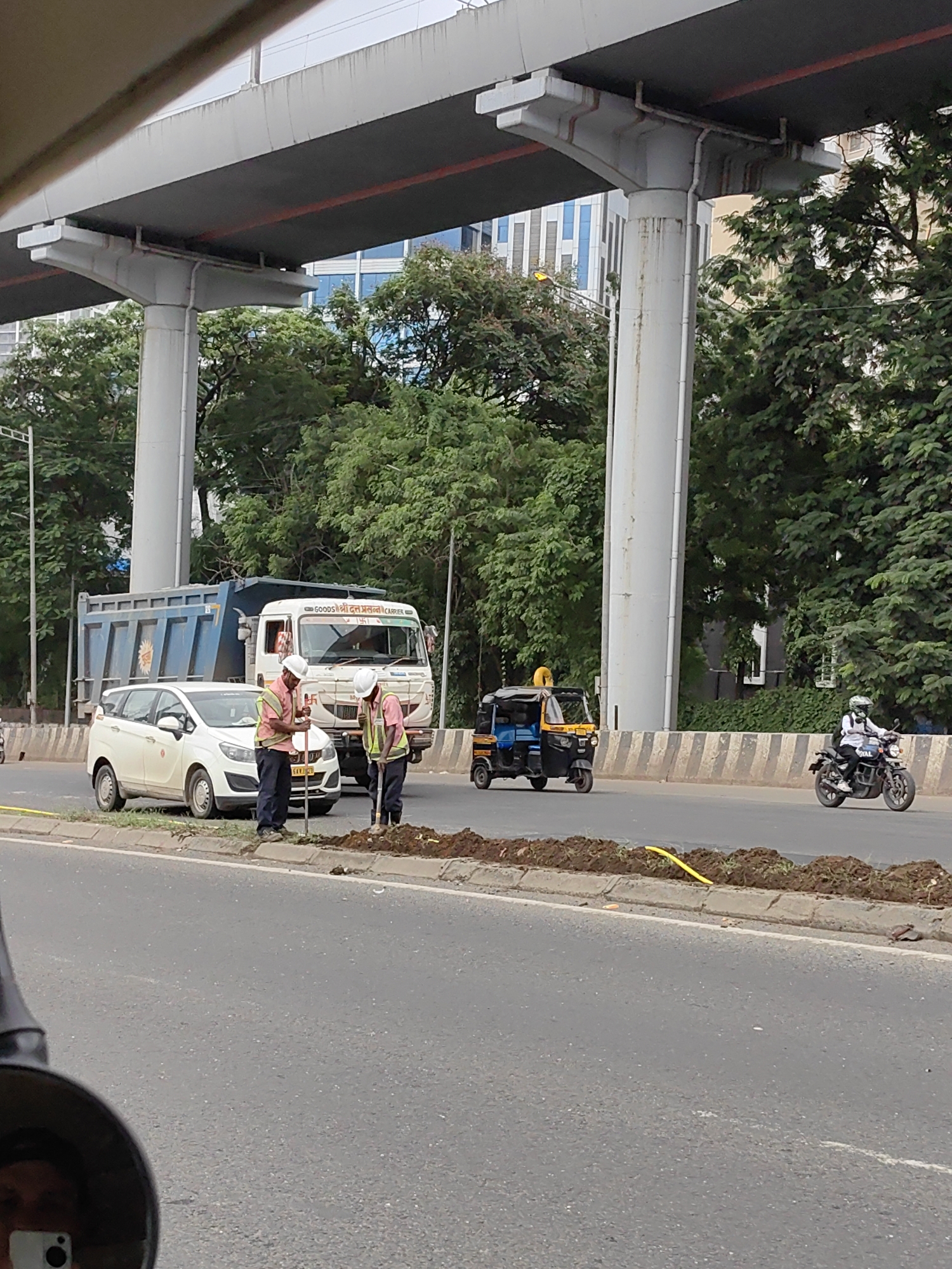 Two construction workers in reflective vests and hard hats are working on the side of a road, digging into a mound of dirt. A white car is parked behind them, and a blue dump truck is further behind the car. An auto-rickshaw and a motorcycle are driving on the road. In the background, there is a large elevated highway supported by concrete pillars and lush green trees. The scene appears to be in Mumbai, India, given the presence of the auto-rickshaw and the style of the vehicles. The overall environment suggests a busy urban setting undergoing some sort of road or infrastructure work.