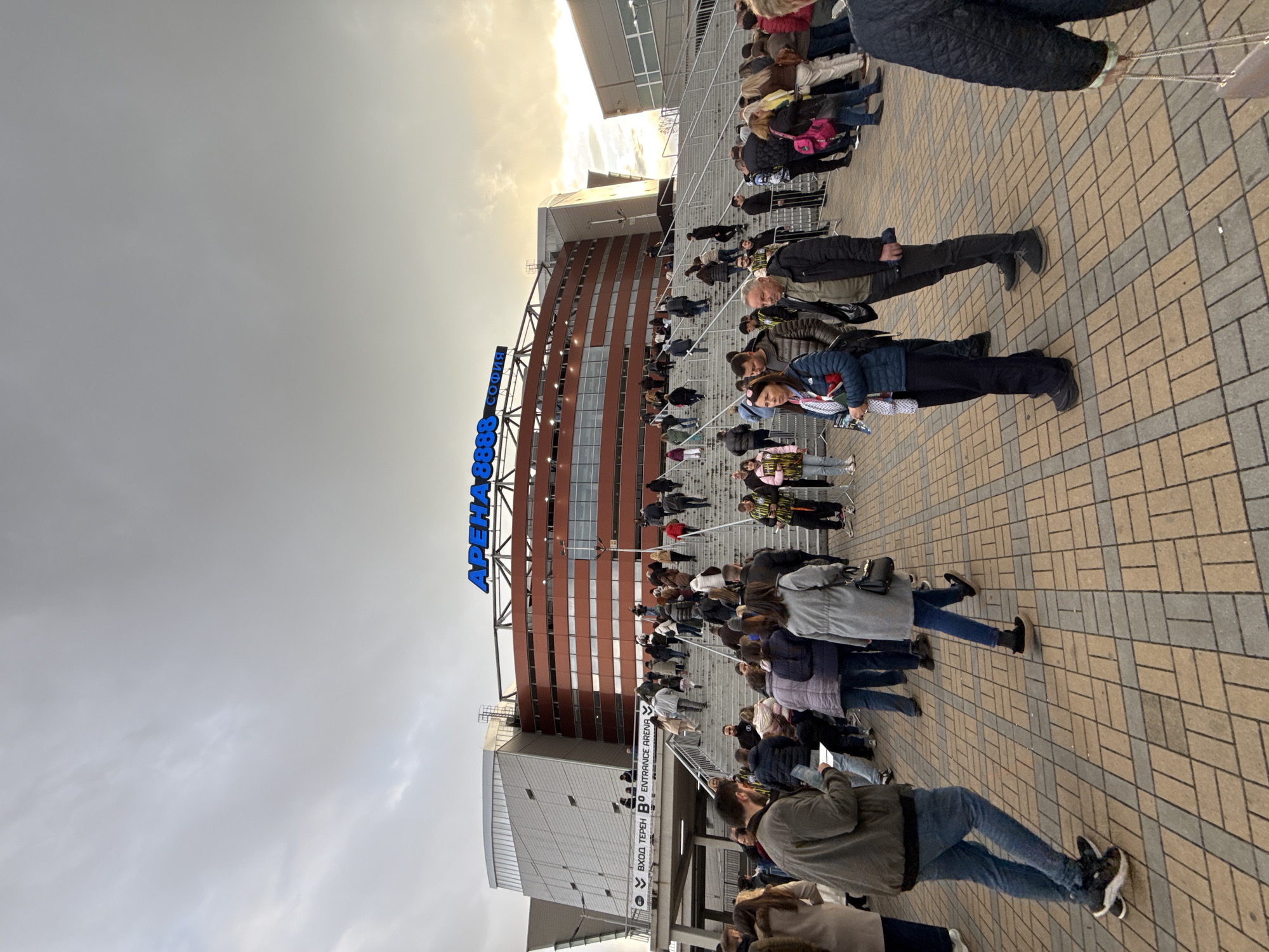 A crowd of people are standing on a paved area and on stairs leading up to a large building with the words "APEHA COOMA" in blue lettering near the top. The building has a facade with horizontal bands of brown and orange. Signage indicates an entrance. The sky is overcast. The location is likely the Arena Sofia in Sofia, Bulgaria.