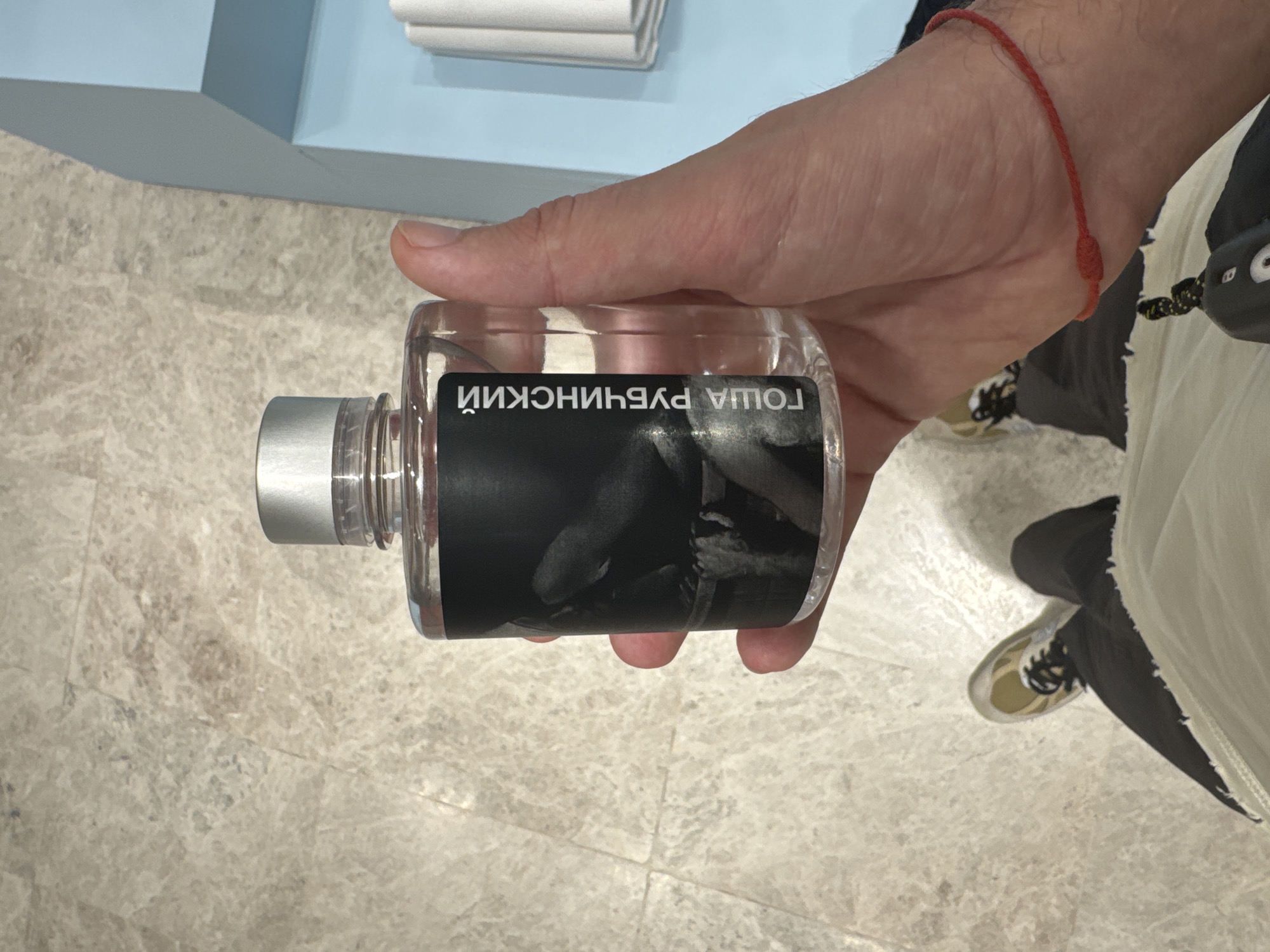 A person's hand holds a clear bottle of perfume with a black label featuring a grayscale image and Cyrillic text. The text on the label reads "ГОША РУБЧИНСКИЙ". The person is wearing dark pants and light-colored sneakers with yellow accents. A thin red string bracelet is visible on the person's wrist. The background appears to be a tiled floor and a pale blue structure. No specific location in Moscow, Russia, can be determined from the image.