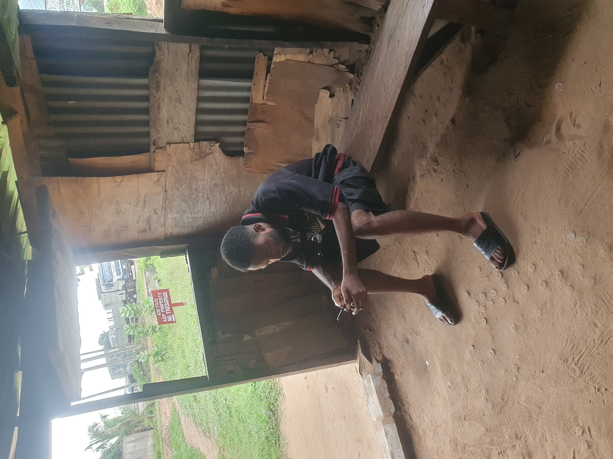 A Black male is sitting on a wooden bench in what appears to be a dilapidated, open-sided structure. He is wearing a dark polo shirt with red trim and dark shorts, and black slide sandals. He is looking down at a smartphone he is holding in his hands. The ground inside the structure is sandy dirt, and there are several small, round objects scattered on the ground, possibly coins or pebbles. Outside the structure, there is a grassy area with some trees and a dirt path. In the background, there are large trucks visible, suggesting an industrial or transport area. A red sign with white lettering reads "KOGI STATE ADP PROPERTY NO TRESPASS". The overall impression is a simple, utilitarian outdoor space, possibly a waiting area or a small shelter, located in Lokoja, Nigeria.