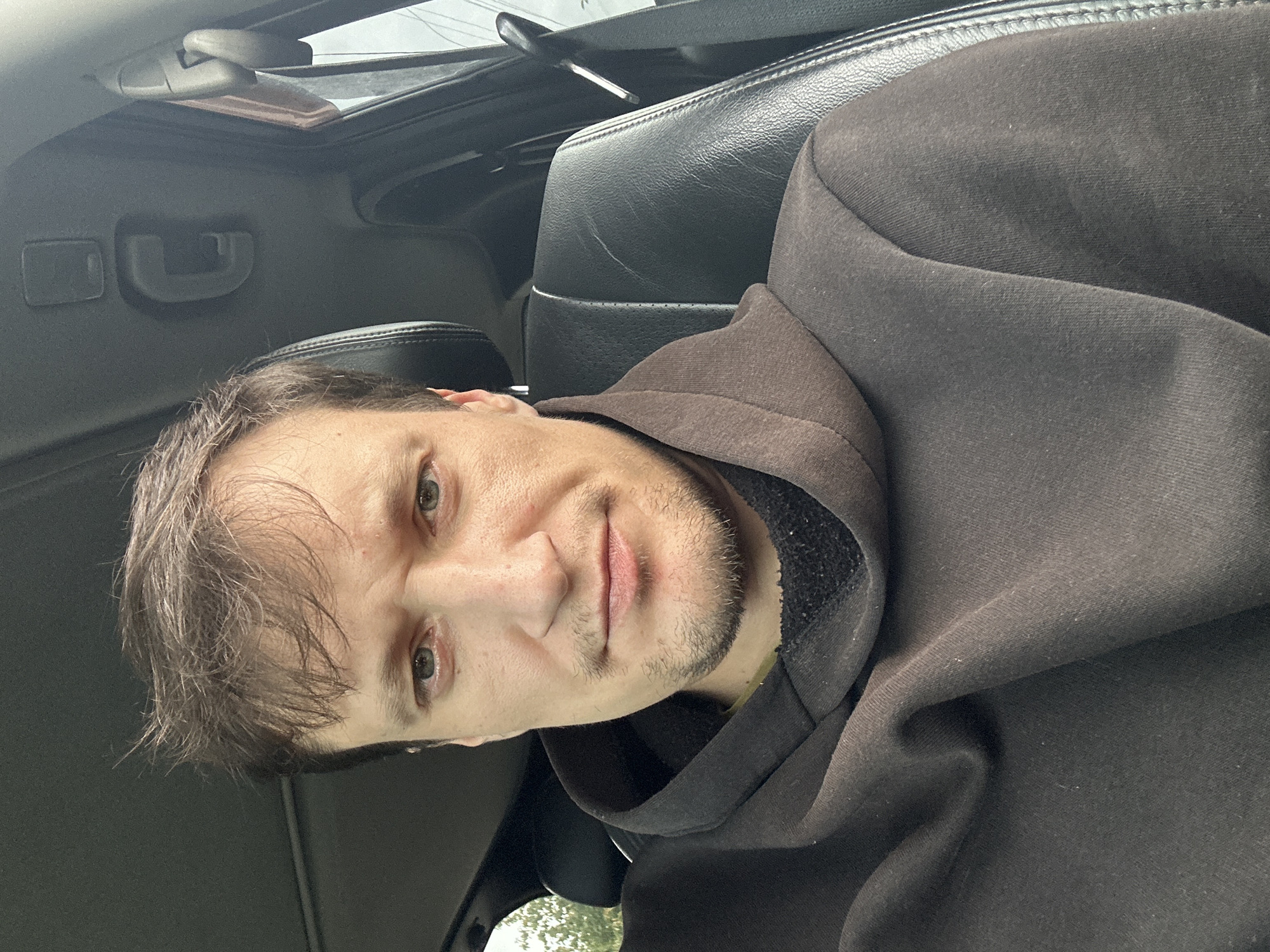A man is sitting in the driver's seat of a car, looking directly at the camera. He has light brown hair, light blue eyes, and a slight stubble on his face. He is wearing a dark brown hooded sweatshirt. The interior of the car is visible, including the black leather seat, the ceiling with a grab handle, and the window with trees outside. The lighting suggests it is daytime. The photo was taken in the city of Makiv, Ukraine.