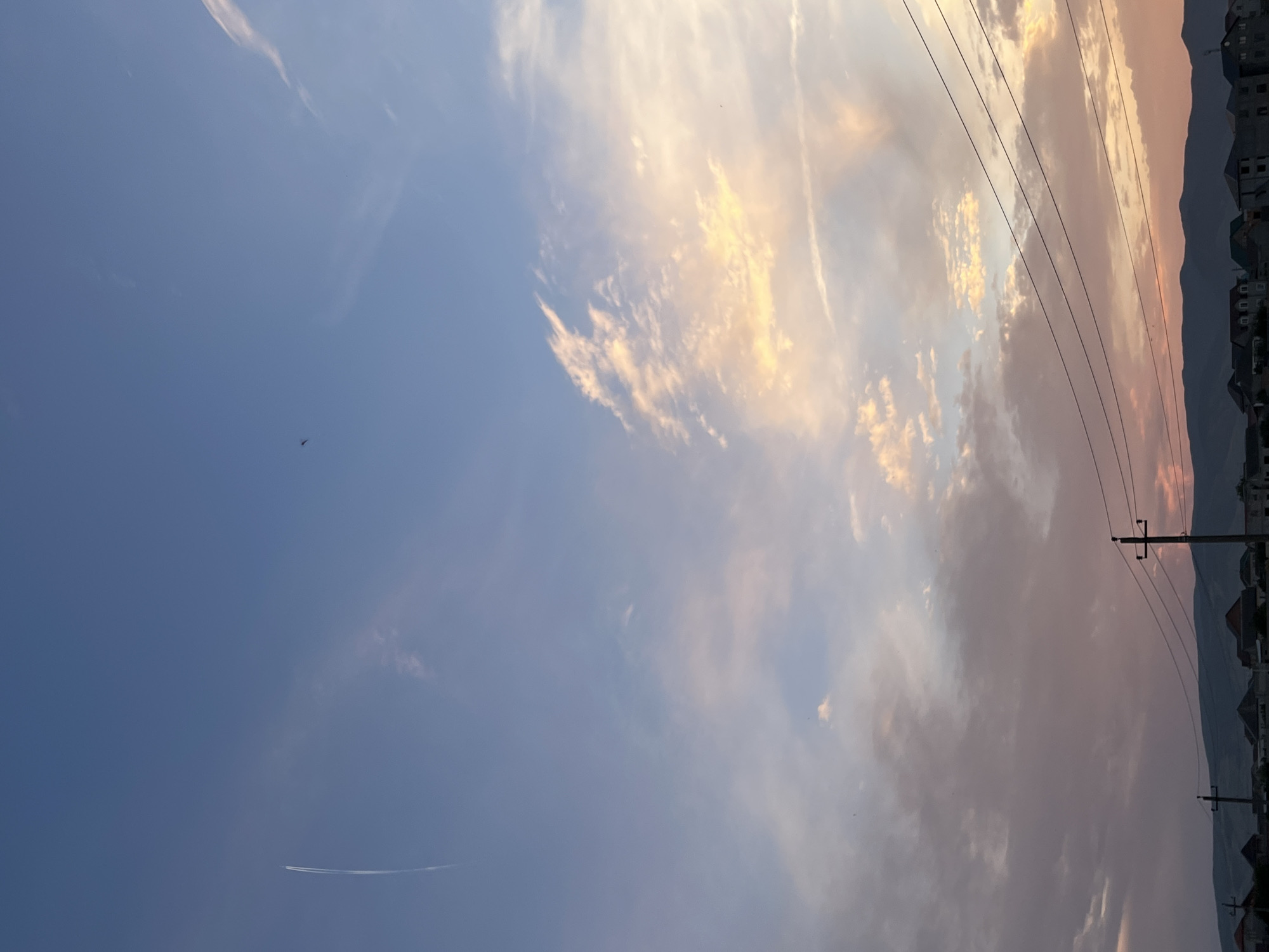 The image captures a twilight sky over the city of Kaspiysk, Russia. A faint contrail from an aircraft is visible in the upper left quadrant, and a distant bird can be seen in the upper right. The sky transitions from a clear blue at the top to soft hues of pink, orange, and purple clouds towards the horizon, indicating the setting or rising sun. In the foreground, silhouetted against the colorful sky, are the outlines of buildings and houses, characteristic of a residential area. Power lines and utility poles are also prominent features, running horizontally across the mid-ground. A mountain range forms the backdrop to the city. No people are visible in the image.