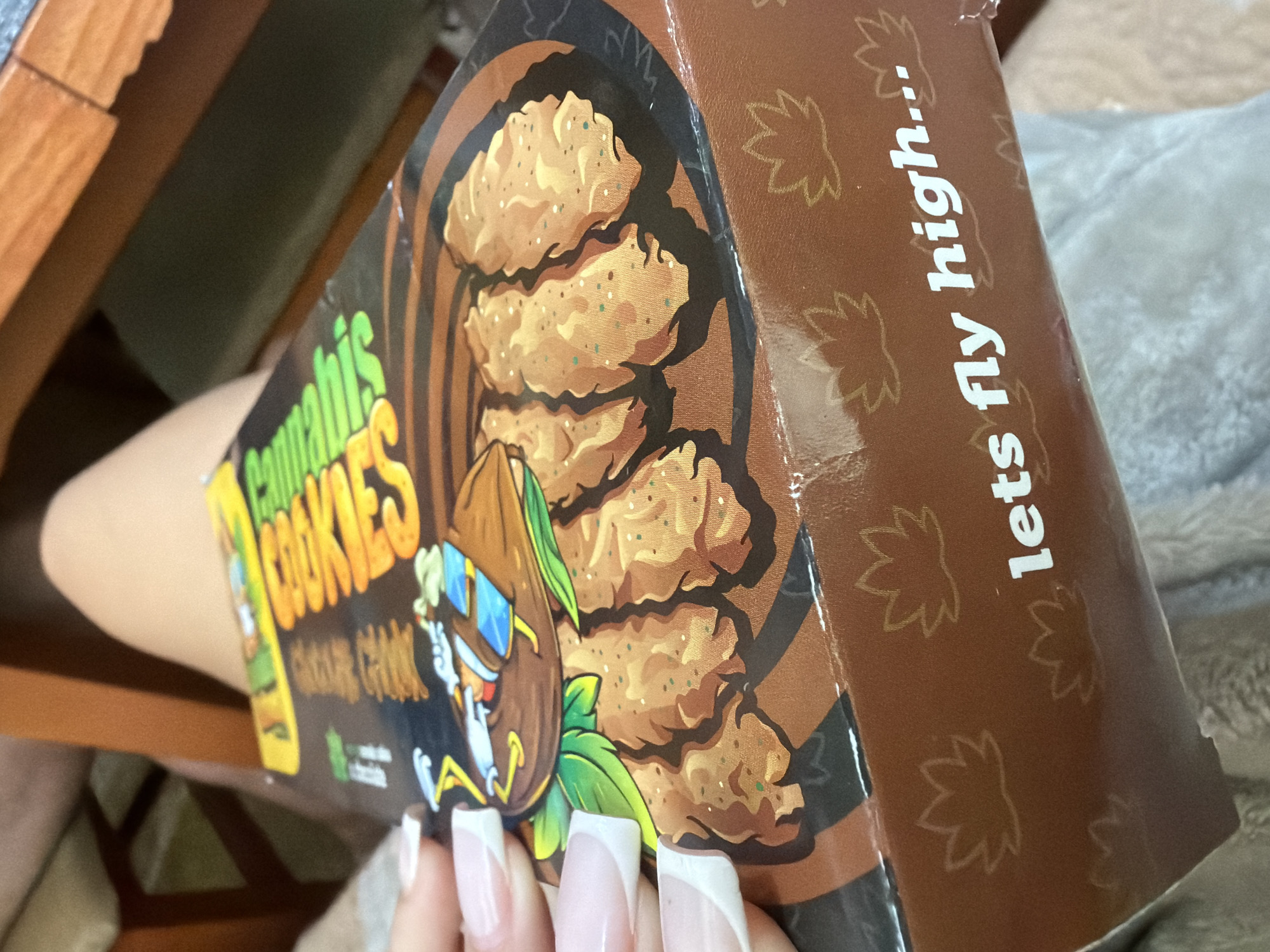 A close-up, slightly overhead shot features a person's hand with white French-tipped nails holding a brown cardboard box. The box is clearly branded with the words "Cannabis COOKIES" in green and yellow lettering. Below "COOKIES," smaller, partially obscured text appears, possibly including "CBD." A small green square symbol, potentially indicating a dietary claim like vegan or plant-based, is also visible.
The front of the box displays an illustration of several golden-brown cookies with speckles, resembling chocolate chip or oatmeal varieties. Beside the cookies, a cartoon character, possibly a bull or cow, is depicted wearing a hat and sunglasses. On the side panel of the box, the phrase "lets fly high..." is written in white, uppercase text against a brown background, which is subtly patterned with outlined cannabis leaf shapes.
The person's hand is in the foreground, specifically in the lower right portion of the image, grasping the box. In the blurred background, wooden furniture, possibly a table or chair armrest, is visible on the left, while a soft, light-colored blanket or fabric can be seen on the right. The scene appears to be indoors. The primary activity captured is the holding or presentation of the product box. The location context provided for this image is Burgas, Bulgaria. No specific time of day, weather, or discernible emotions are conveyed by the image.