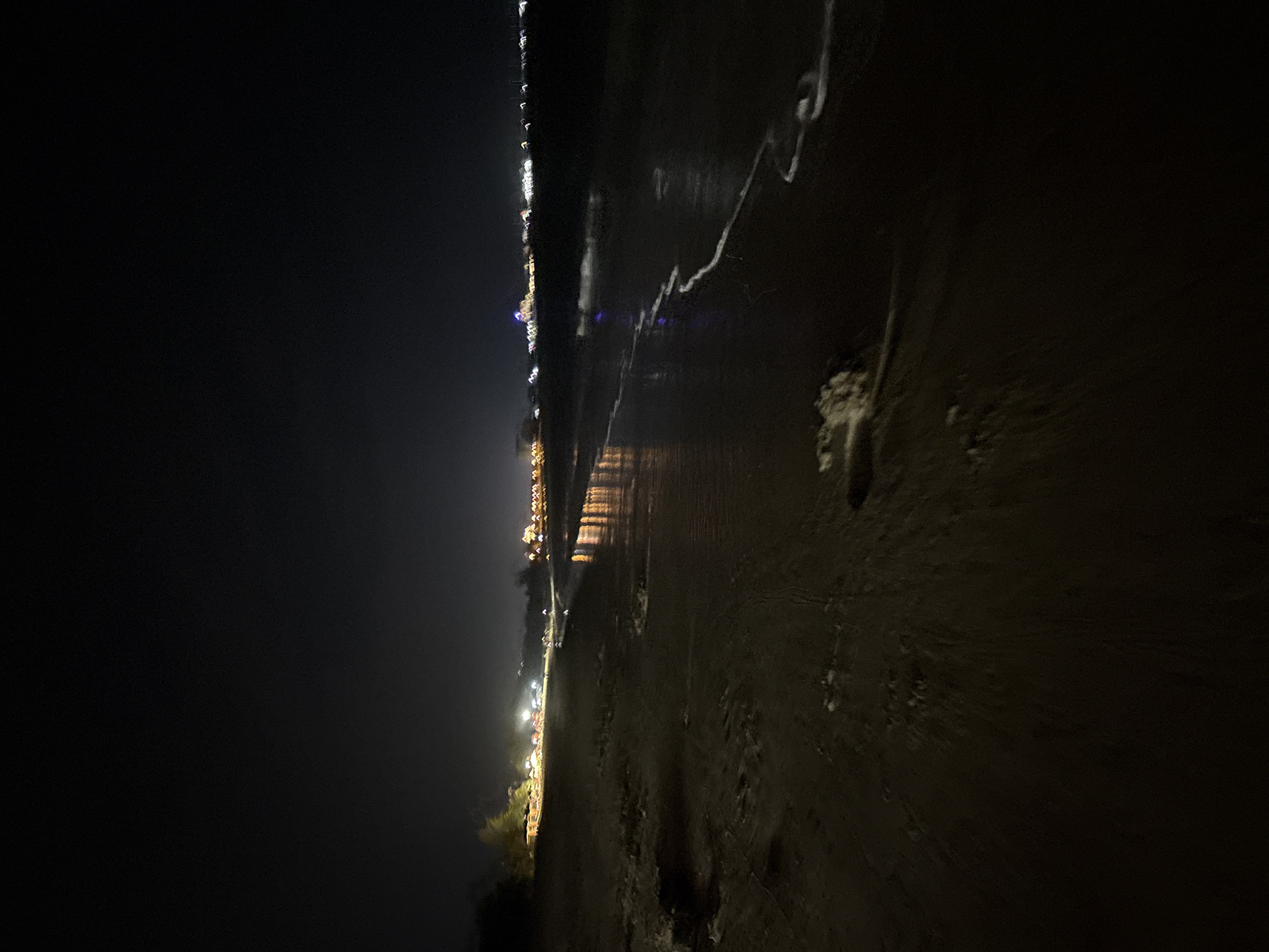 This media file captures a nighttime coastal scene. The image depicts a wide expanse of dark night sky on the left, transitioning to a body of water and a sandy or muddy shoreline on the right. Along the distant horizon, a line of artificial lights from a town or settlement illuminates the far coast, casting faint reflections on the water's surface. White, wavy lines of what appear to be sea foam or gentle waves are visible in the water closer to the foreground. The immediate foreground consists of a dark, textured beach, possibly sand or mud, with subtle variations in its surface. There are no discernible people or specific objects in the immediate foreground, and no activity is taking place. The time of day is clearly night, and the weather appears calm, allowing for clear visibility of the distant lights. The overall mood is one of tranquility and solitude. No visible text is present in the image. The location is consistent with Dong Hai Ward, Vietnam, known for its coastal features.