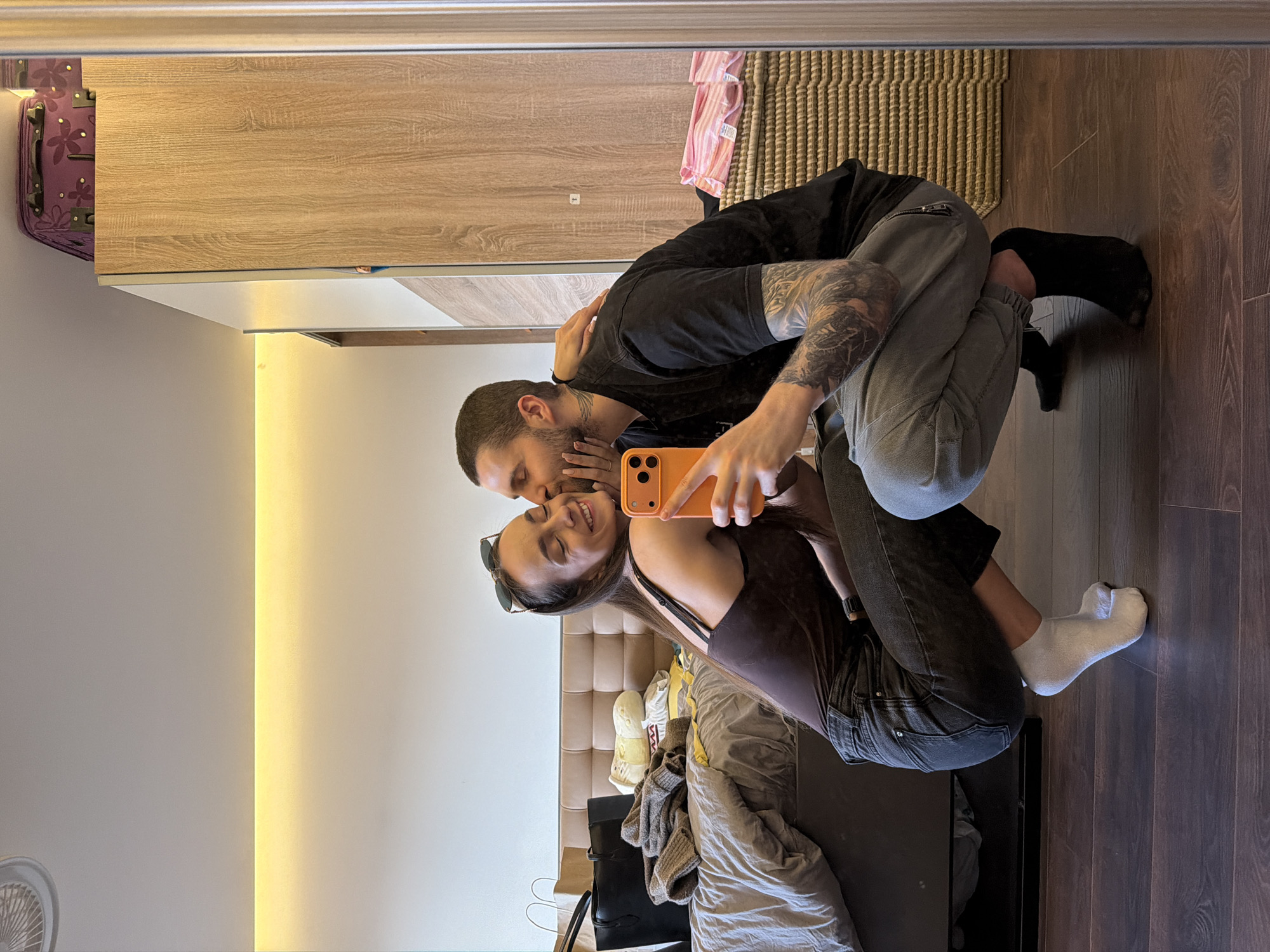 A couple is taking a selfie in front of a mirror. The woman is in front wearing a brown tank top and dark jeans, and a white sock on her right foot. The man is behind her, wearing a black t-shirt and grey sweatpants, and a black sock on his left foot. He is holding a bright orange phone. The background includes a wooden closet, a wicker basket, a bed with a beige headboard, and a room with a white wall and wooden floor. The image shows a reflection, suggesting an indoor location. There is no information available in the picture to suggest this scene is in Sofia, Bulgaria.