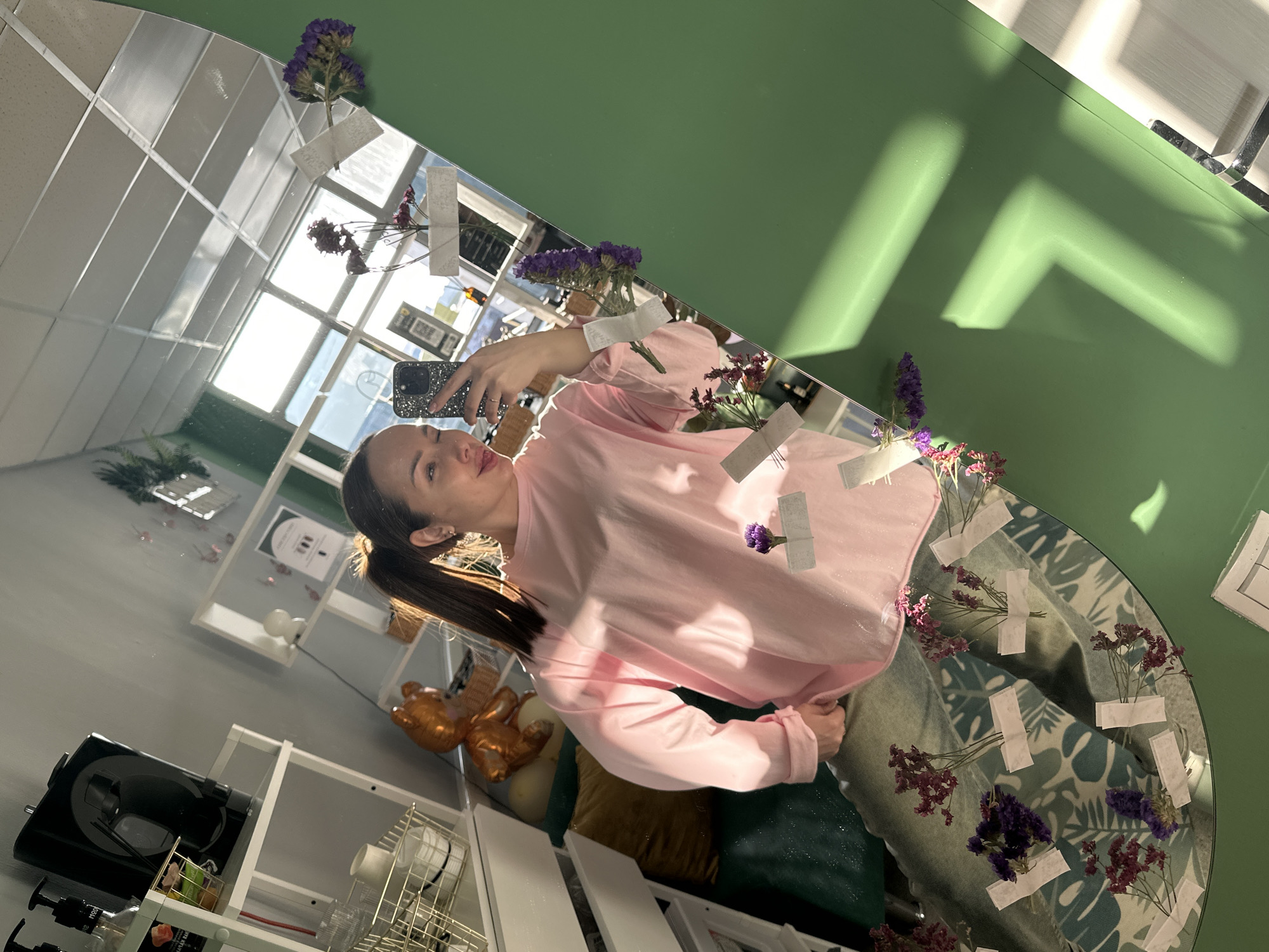 A female individual is depicted taking a mirror selfie within a commercial establishment in Chaikovsky, Russia. The subject, with brown hair tied in a high ponytail, wears a loose-fitting light pink long-sleeved shirt and light-colored patterned pants. She holds a smartphone with a dark, glittery case in her right hand, positioned to capture her reflection. Her expression is a slight smile with a partially closed left eye.
The selfie is taken in a large, irregularly shaped mirror featuring a wide, matte green border. Several bundles of dried purple and reddish-brown flowers are affixed to the mirror's surface using small white adhesive strips.
The reflection reveals a well-lit interior with ample natural light entering through large windows, which also reflect a white grid-panel ceiling. On the left side of the reflection, white open-frame shelving units display various items, including bottles with pump dispensers, a black electronic device, and gold-colored wire baskets containing white ceramic ware. A shiny, gold-colored inflatable bear balloon is visible behind the subject's left shoulder, resting on a light-colored object. A dark green upholstered seating area with a gold throw pillow is partially reflected below the subject's waist. Light gray walls are visible in the background, along with a reflected QR code or sign. Strong sunlight casts distinct shadows onto the green mirror border.