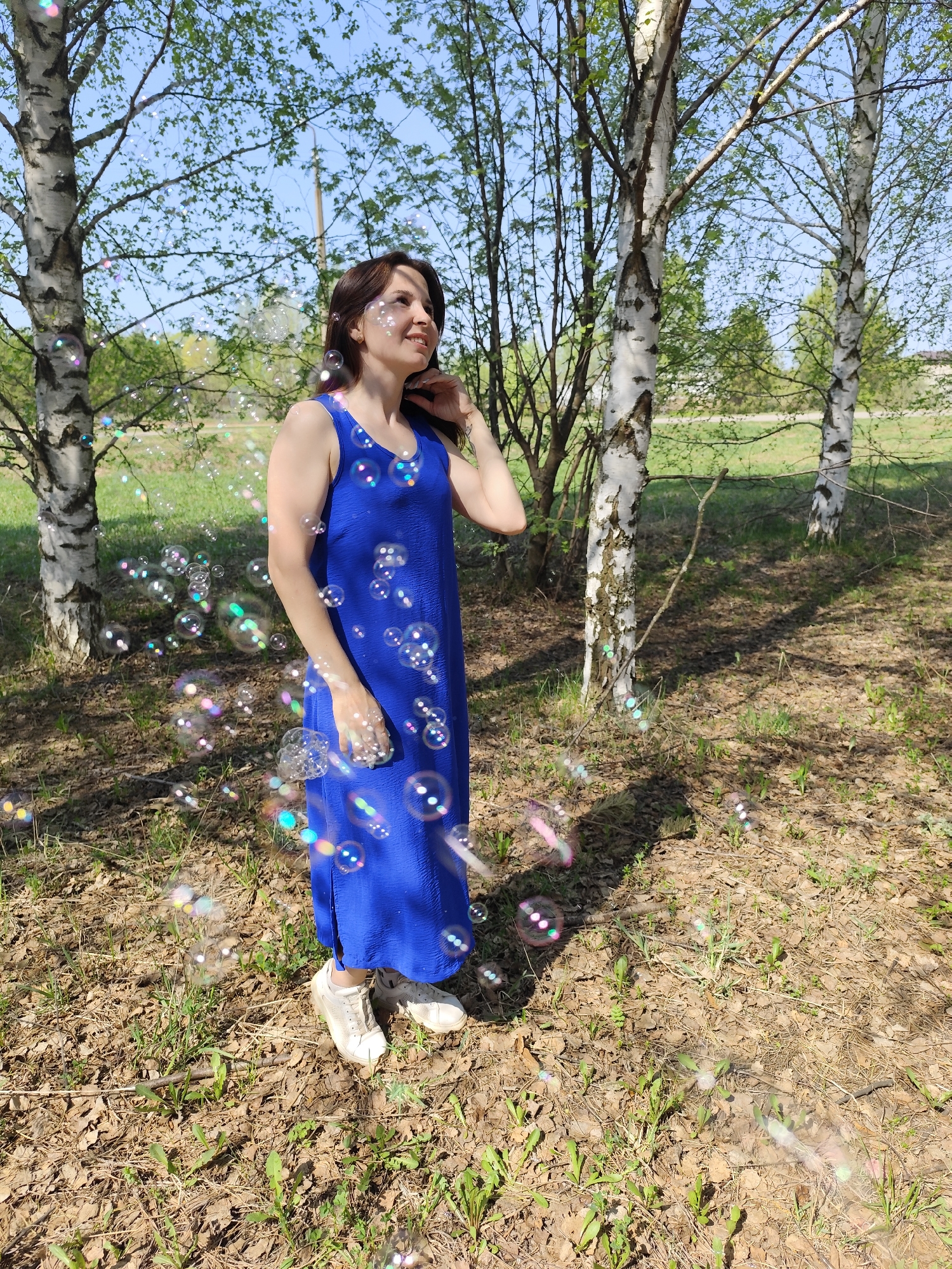 A woman in a blue dress stands among birch trees. Bubbles float around her. The ground is covered in fallen leaves and sparse grass. The location is Фоки, Russia.