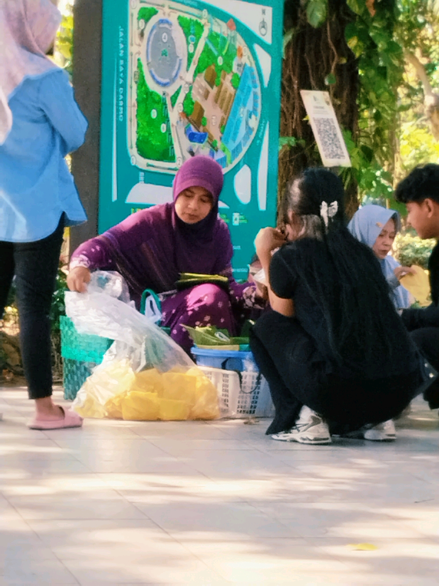 In Surabaya, Indonesia, at the Surabaya Zoo (Kebun Binatang Surabaya), a woman in a purple hijab and dress is selling food items to visitors. She is seated near a large, colorful map of the zoo and a tree with a QR code attached to it. In front of her, there are baskets of food wrapped in yellow plastic. Several other people, including a woman in a blue hijab and a young boy, are also present, some of whom appear to be purchasing food. The scene takes place outdoors, with sunlight casting shadows on the tiled ground.
