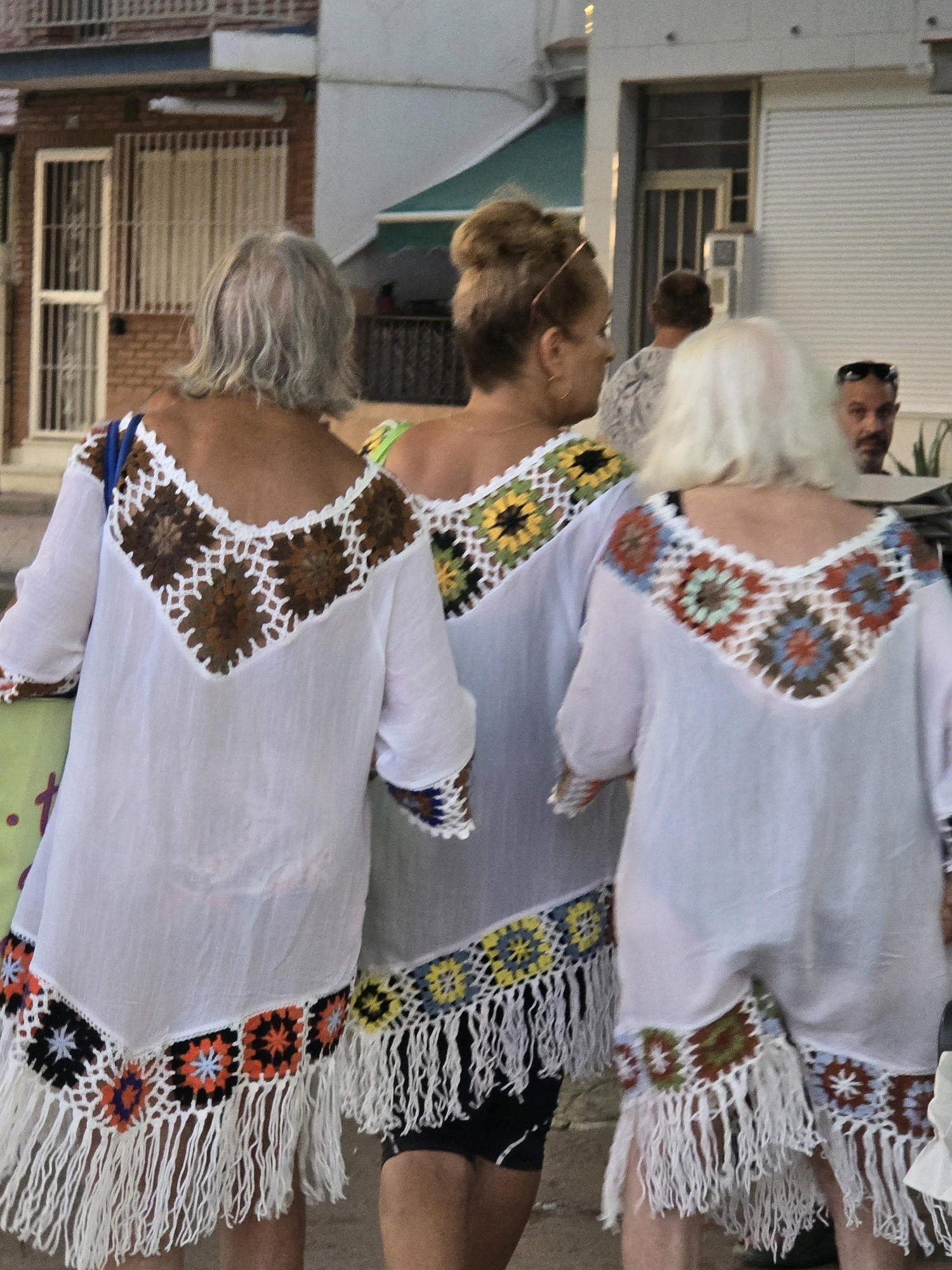 The image shows three women walking away from the camera. The woman on the left has grey hair and is wearing a white tunic with a crocheted V-neckline in brown and white. The woman in the middle has her hair up in a bun and is wearing a white tunic with a crocheted V-neckline featuring yellow, green, and white floral patterns. The woman on the right has blonde hair and is wearing a white tunic with a crocheted V-neckline in red, blue, green, and white geometric patterns. All three tunics have fringed hemlines. They appear to be walking in a pedestrian area, possibly a street or plaza, with buildings in the background. The buildings have brick and white facades, and one has a window with vertical bars. The overall atmosphere suggests a warm climate, consistent with the city of Málaga, Spain.