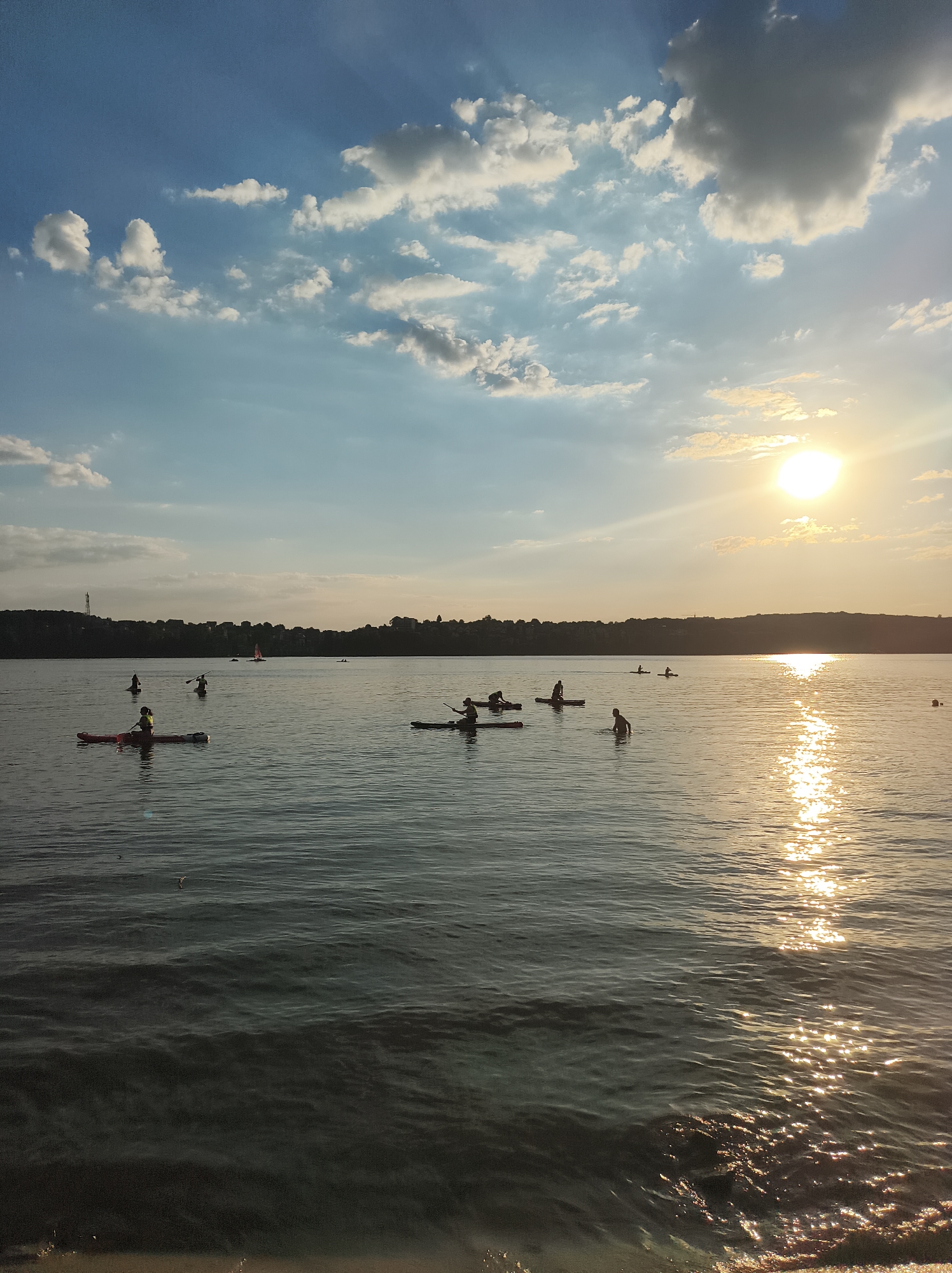 The image depicts Ternopil Lake in Ternopil, Ukraine, during sunset. The sun is setting on the right side of the frame, casting a warm, golden glow on the water and illuminating a path of reflection. Several individuals are engaged in water activities, primarily kayaking and paddleboarding, scattered across the lake. A silhouetted treeline with buildings and a prominent telecommunications tower is visible in the background, marking the edge of the city. The sky is a mix of blue with scattered white and gray clouds, and some rays of sunlight are visible breaking through. The foreground shows the rippling surface of the lake with small waves.