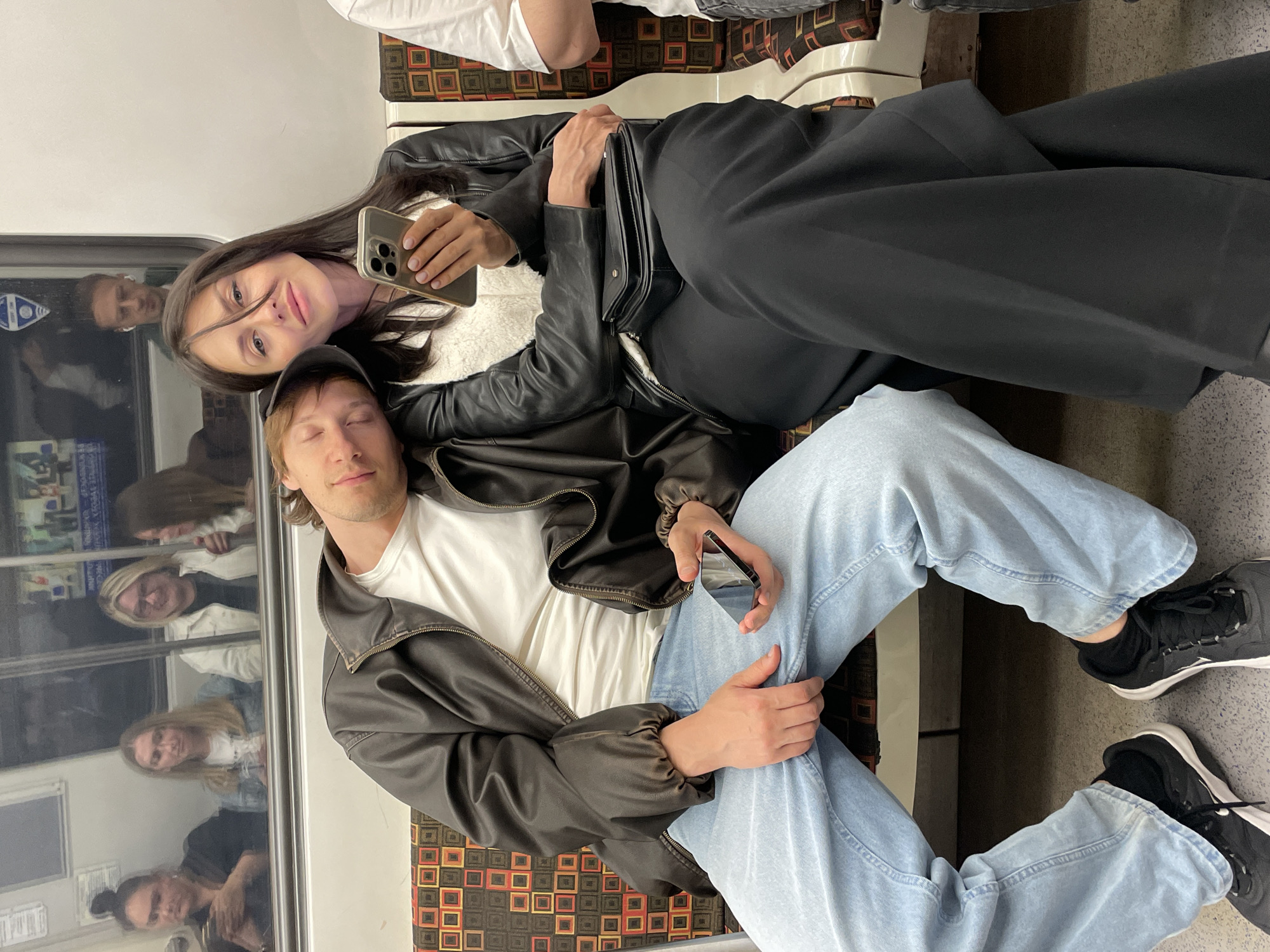A candid medium shot captures two individuals, a man and a woman, seated next to each other on a subway train, their bodies leaning towards one another. The man, on the left, appears to be asleep, his head resting on the woman's shoulder, and he is wearing a dark brown leather jacket over a white t-shirt and light blue jeans. His eyes are closed, and a black baseball cap is perched on his head. In his right hand, he holds a dark-colored smartphone.
The woman, on the right, is awake and looking towards the viewer, holding a gold smartphone in her right hand. She is dressed in a black leather jacket over a white fuzzy top and black wide-leg trousers. Her head is turned slightly, and her gaze is direct, with her lips slightly parted. A black purse is placed on her lap, just above her trousers.
The subway car's interior is visible, with patterned seating in shades of orange, brown, and cream. The background shows reflections in the window, revealing the faces of other passengers and glimpses of the subway tunnel. The lighting is dim, characteristic of an indoor urban transit environment. The overall impression is one of intimacy and quiet repose amidst the hustle of city travel.