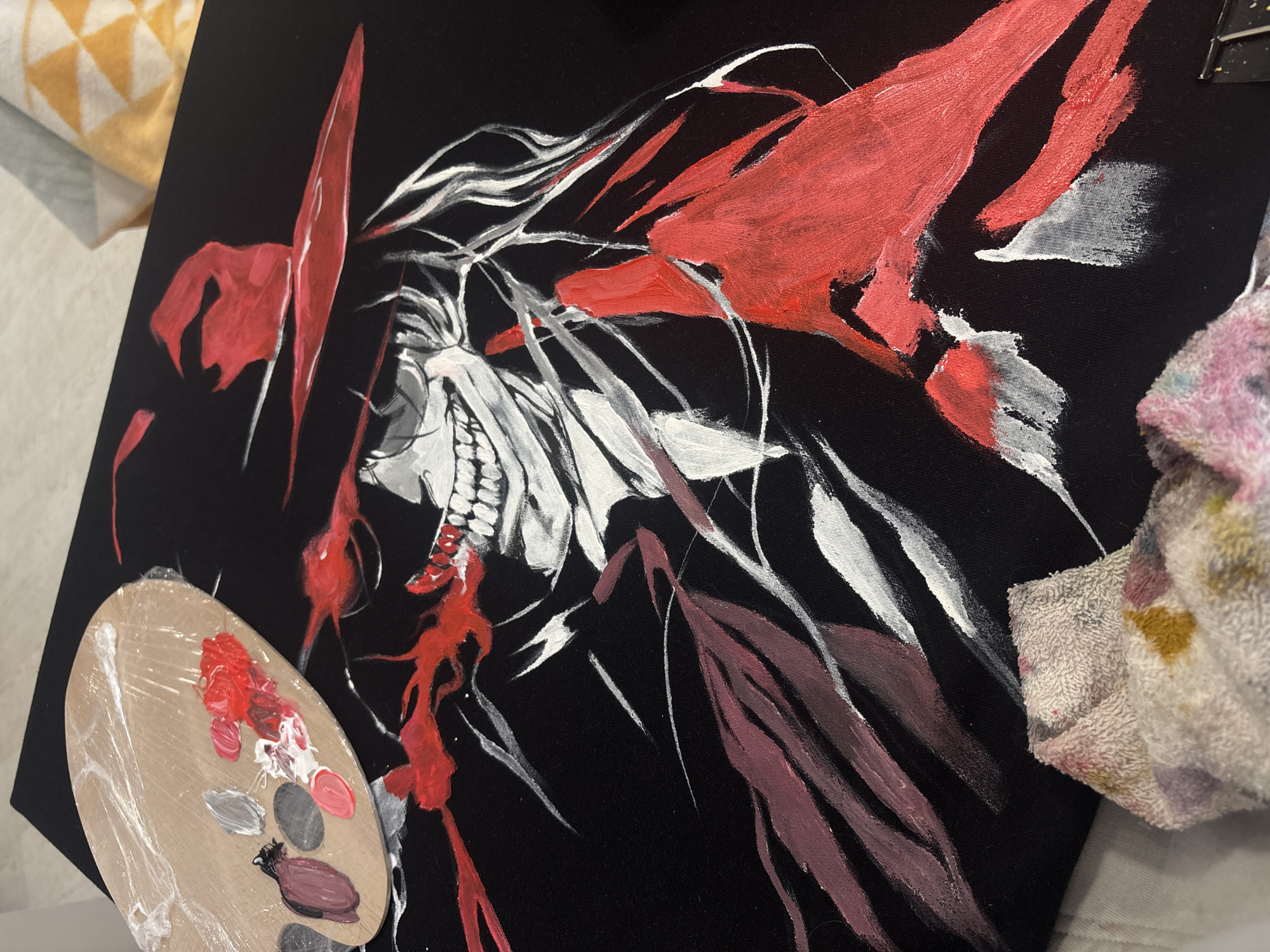 The image shows a black canvas painting in progress. The painting depicts a stylized figure with a skull-like face, sharp teeth, and white hair or flowing forms. Red and maroon paint is used for additional features around the head. A wooden paint palette with various colors including red, white, gray, and brown sits to the left of the canvas. A multicolored cleaning cloth is located to the right of the canvas. A cushion is partially visible in the upper left corner of the frame. The image does not contain sufficient information to determine the location in Krasnoyarsk, Russia.