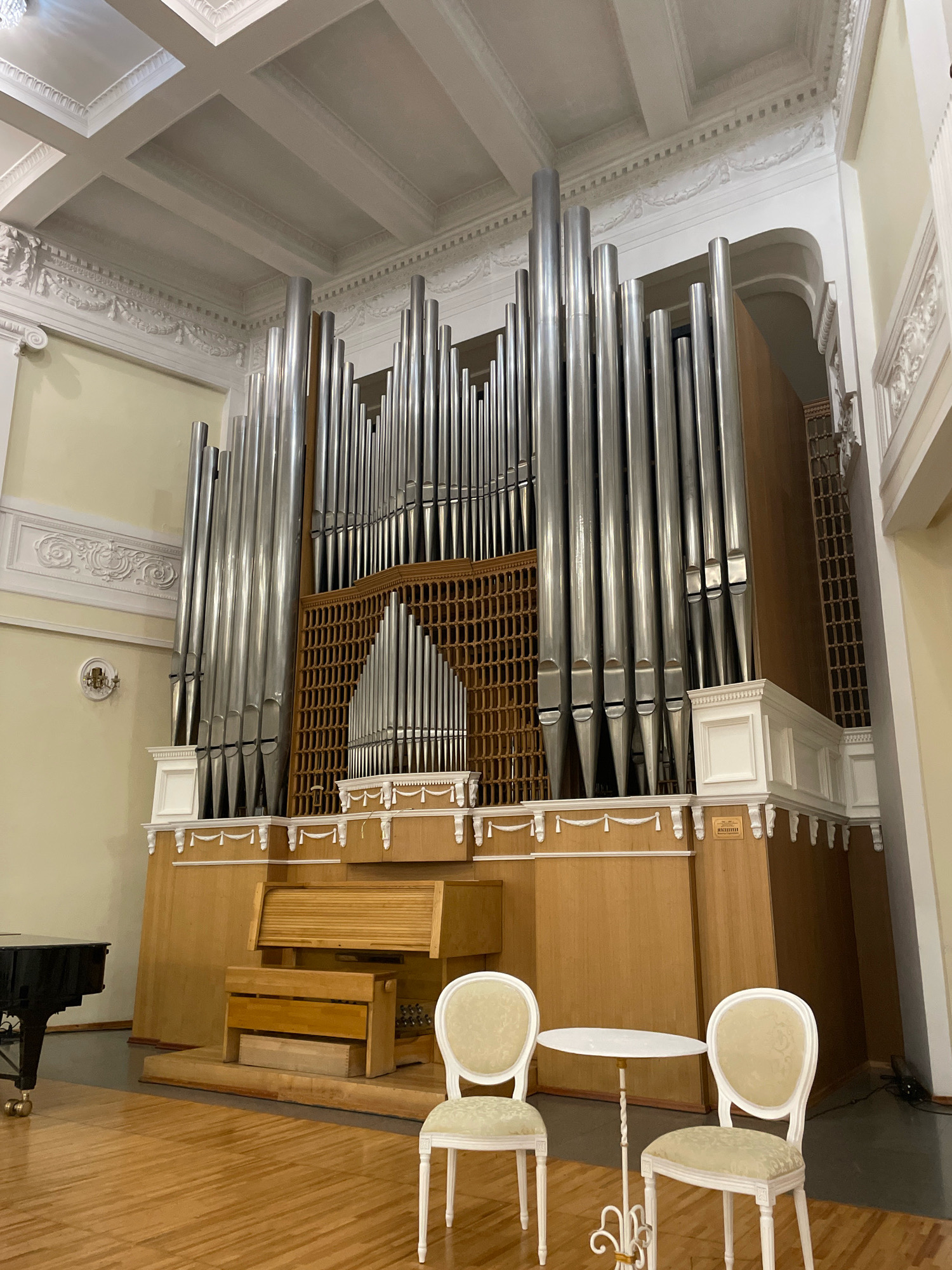 The image depicts a large pipe organ in what appears to be a concert hall or church in Omsk, Russia. The organ is the central focus, with numerous tall, polished metal pipes dominating the upper portion of the scene. Below the pipes, wooden elements form the console and a decorative lattice. In the foreground, two white chairs with ornate carved legs and patterned upholstery flank a small, round white table. To the left, a portion of a black grand piano is visible, its surface reflecting the ambient light. The flooring is a polished wooden parquet. The ceiling above features intricate plasterwork and coffered panels. The walls are a pale yellow and adorned with decorative moldings. The overall impression is one of a grand, well-maintained interior, likely a venue for musical performances.