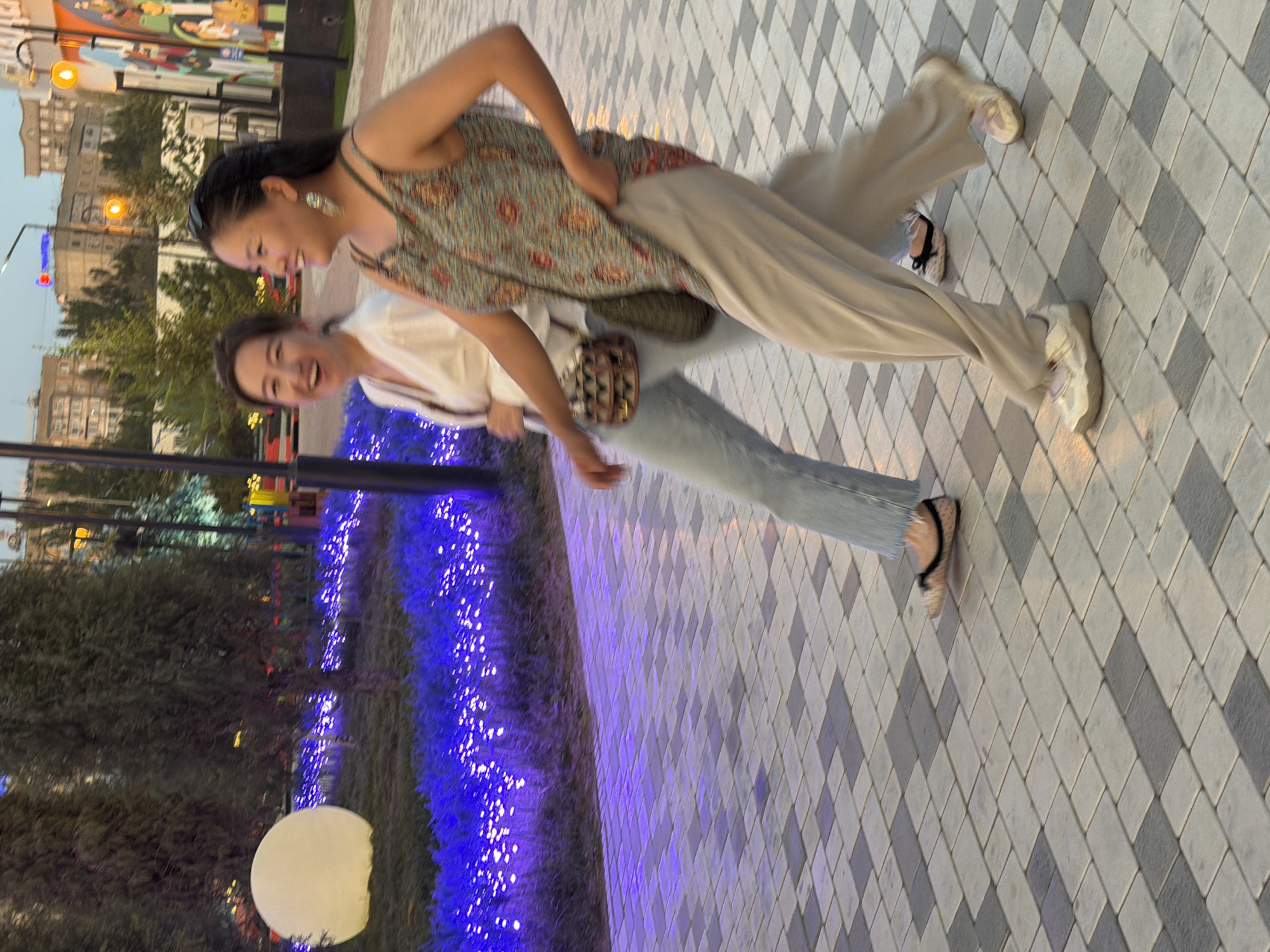 Two women are walking on a paved walkway in what appears to be a park or public space in the city of Unknown, Unknown. The woman on the left is wearing a white top and blue jeans, and she is smiling and looking to her right. The woman on the right is wearing a patterned sleeveless top and loose-fitting pants, and she is also smiling and looking to her right. She has large earrings and is wearing sunglasses on her head. The background includes trees, blue lights, a large white sphere, and buildings in the distance. The walkway is made of gray and white rectangular tiles. The lighting suggests it is late afternoon or early evening.