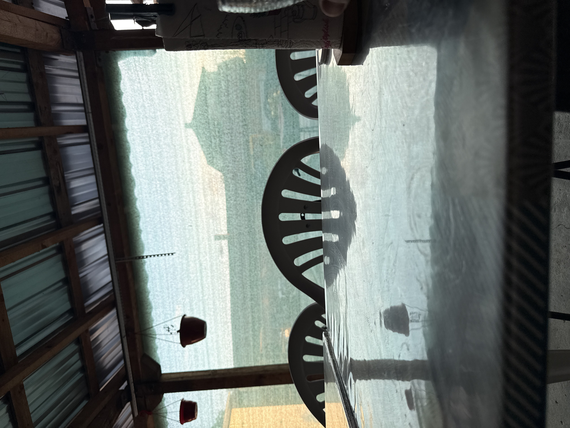 The image depicts an outdoor scene, likely taken from a covered patio or porch. Visible is the interior of a wooden structure with a corrugated metal roof. Hanging flower pots are suspended from the beams. Through a partially obscured screen, a landscape featuring a body of water, a shoreline, and a prominent hill or structure in the distance is visible. Inside the structure, a table with a reflective surface is present, around which are three white, plastic chairs. The scene is Sarnevo, Bulgaria.