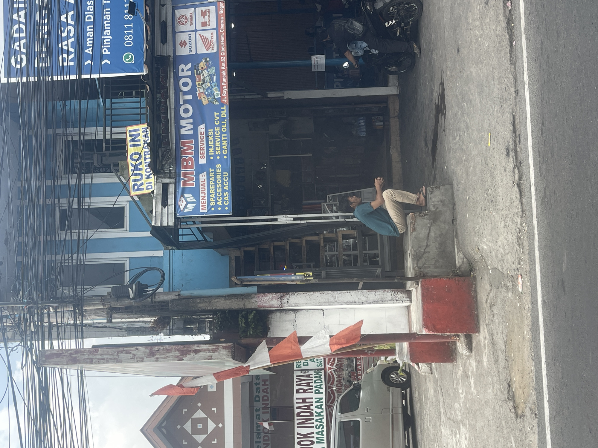 The image depicts a street scene in Cisarua, Indonesia. In the foreground, a grey van is parked on the left side of the frame. A man is seated on a concrete step on the right side, facing away from the camera. Behind him, a motorcycle is visible with a person on it. The background features a blue building with various signs. One prominent sign reads "MBM MOTOR" and lists services such as "SPAREPART," "ACCESORIES," and "CAS ACCU," as well as "SERVICE : INJEKSI," "SERVICE CVT," and "GANTI OLI, DLL." Another sign, partially obscured, indicates "RUOKO INI DI KONTRAKAN." Further up the building, a blue sign with white text is visible, mentioning "GADAI," "RASA," "Aman Dias," and "Pinjaman T." Indonesian flags are also displayed on a pole. The environment appears to be a commercial area with multiple storefronts and overhead electrical wires. The ground is paved with concrete and asphalt, showing signs of wear and a small puddle.