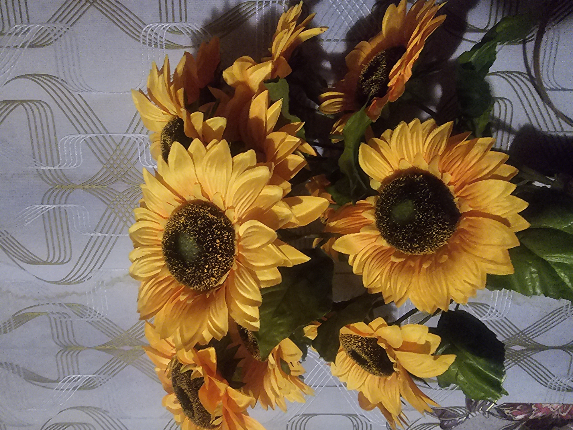 A bouquet of artificial sunflowers is displayed against a patterned wallpaper. The sunflowers are a vibrant yellow with dark centers and green leaves. The wallpaper features a repeating pattern of interwoven, wavy lines in shades of gray and gold. The lighting in the image is somewhat dim, casting shadows on the flowers and wallpaper. The scene is likely an indoor setting, possibly a home or a commercial establishment, and there is no indication of a specific location in Taezhnoye, Russia.