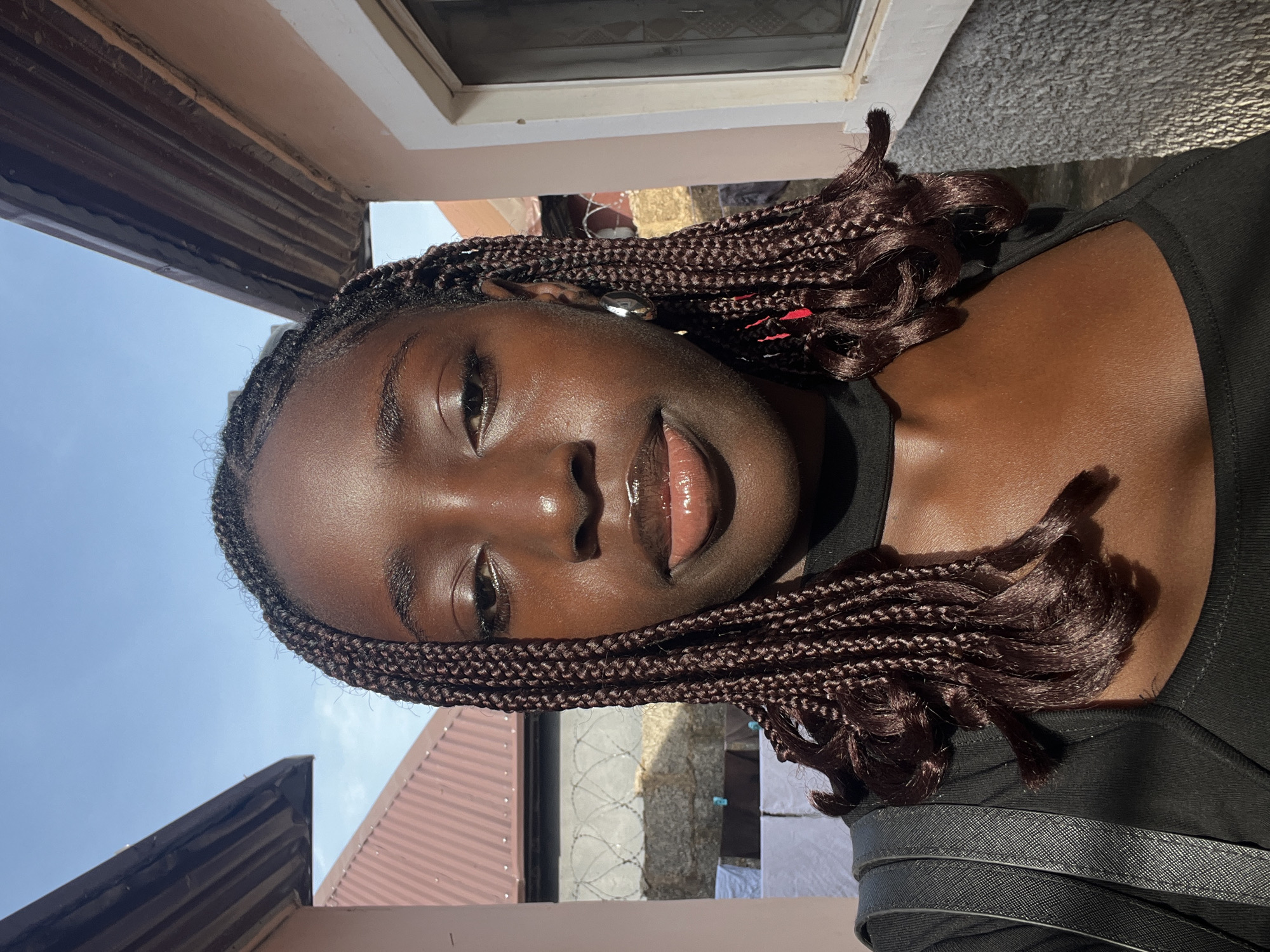 A vertical portrait features a young person with dark skin, angled slightly, looking directly at the viewer with a neutral to subtly pleasant expression. Their lips are glossy, and they have dark braided hair styled in box braids, with some strands resting on their shoulders. A small metallic earring is visible on their right ear. The individual is wearing a black top or shirt, and a black textured bag strap is visible on their right shoulder.
The scene is set outdoors during a clear, sunny day, indicated by the bright, direct sunlight creating highlights on the subject's skin and hair. The background reveals a clear blue sky, suggesting good weather. Architectural elements are visible, including parts of brown corrugated metal roofs, a white window frame, and textured light-colored walls. In the mid-distance, a section of barbed wire fencing and white laundry hanging on a clothesline are discernible. The overall impression is a casual, sunlit portrait in what appears to be a residential or semi-urban outdoor setting. No visible text is present in the image.