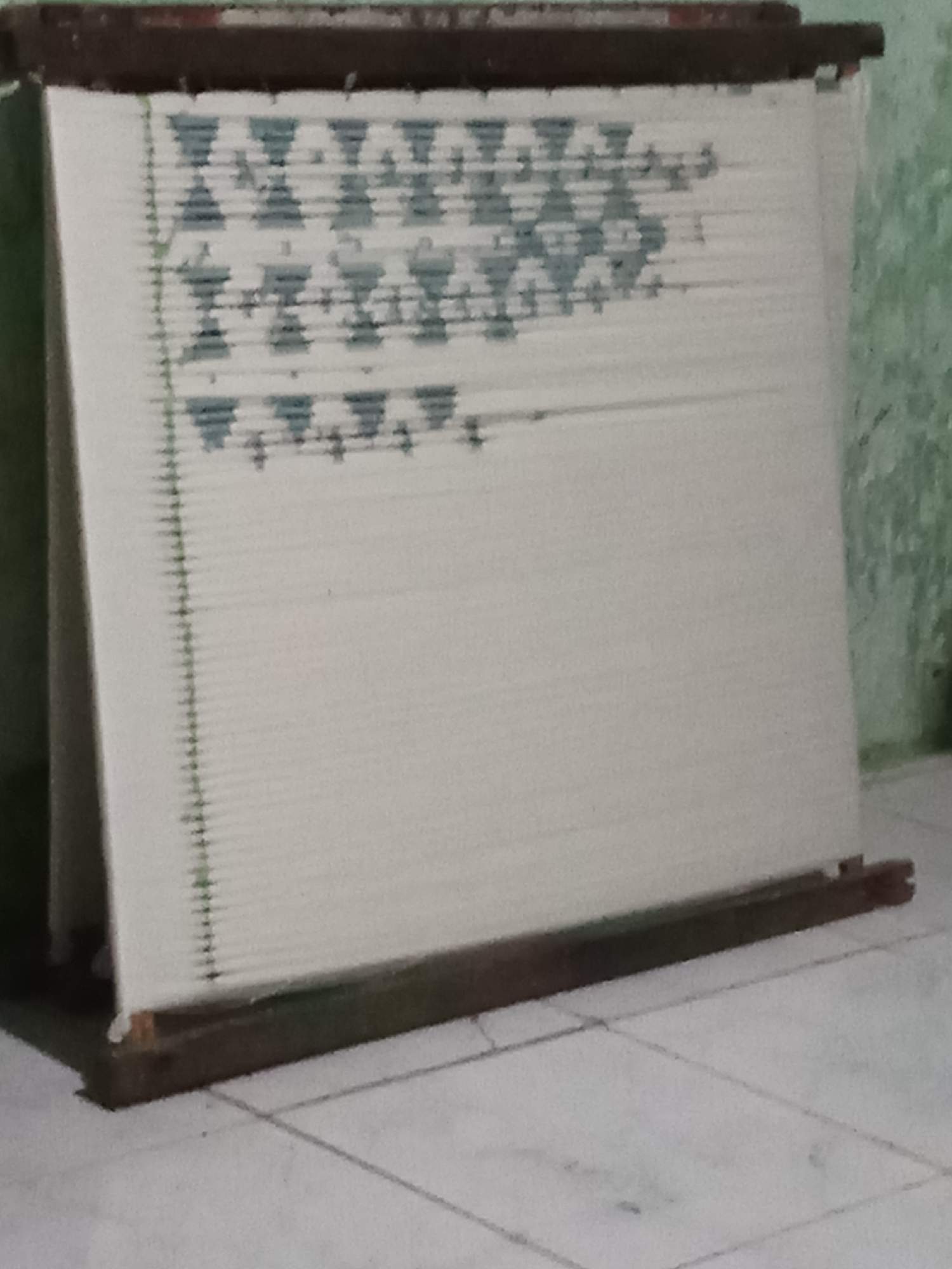 The image shows a loom with a white fabric stretched across it. The fabric has a repeating geometric pattern in a light blue color at the top. The loom itself is made of wood and appears to be old. The background is a blurred green wall and a tiled floor. The scene is lit by natural light. It is not possible to determine the exact location in Pemalang, Indonesia from the image.