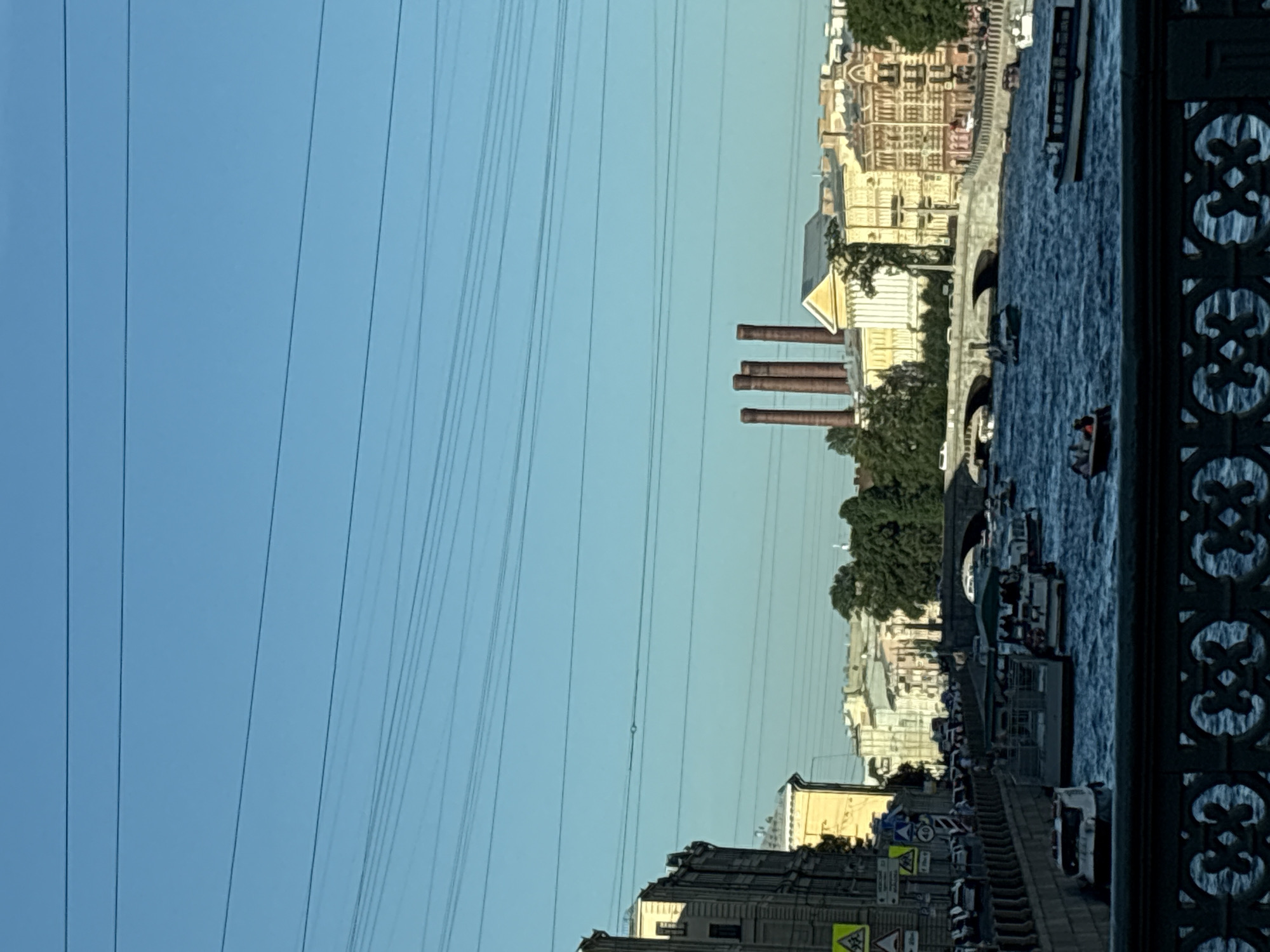 The image shows a view of the Fontanka River in Saint Petersburg, Russia, on a clear day. The river is filled with several boats, including tour boats and smaller vessels. On the left side of the river, there is a street with cars and pedestrians, as well as buildings. In the background, across the river, there are more buildings and a distinctive set of four tall, reddish-brown chimneys. A bridge spans the river in the mid-ground, with traffic visible on it. The sky is a clear blue with several horizontal power lines crisscrossing the upper portion of the image. The foreground features a decorative metal railing.