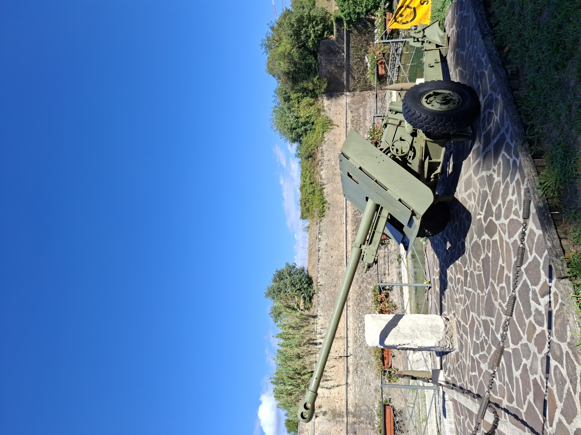 An antique military cannon, painted olive green, is displayed outdoors on a paved area in Peschiera del Garda, Italy. The cannon is positioned on a stone base and has large black wheels. Behind the cannon is a low stone wall covered in greenery, with trees and bushes growing on top. A clear blue sky fills the upper portion of the image. A small body of water is visible beyond the wall, with some decorative planters containing red and pink flowers. A yellow banner with a black emblem is partially visible to the right of the cannon. A concrete pillar stands to the left of the cannon, and metal chains with cylindrical links are draped on the ground in the foreground. The scene appears to be part of a historical exhibition or memorial.