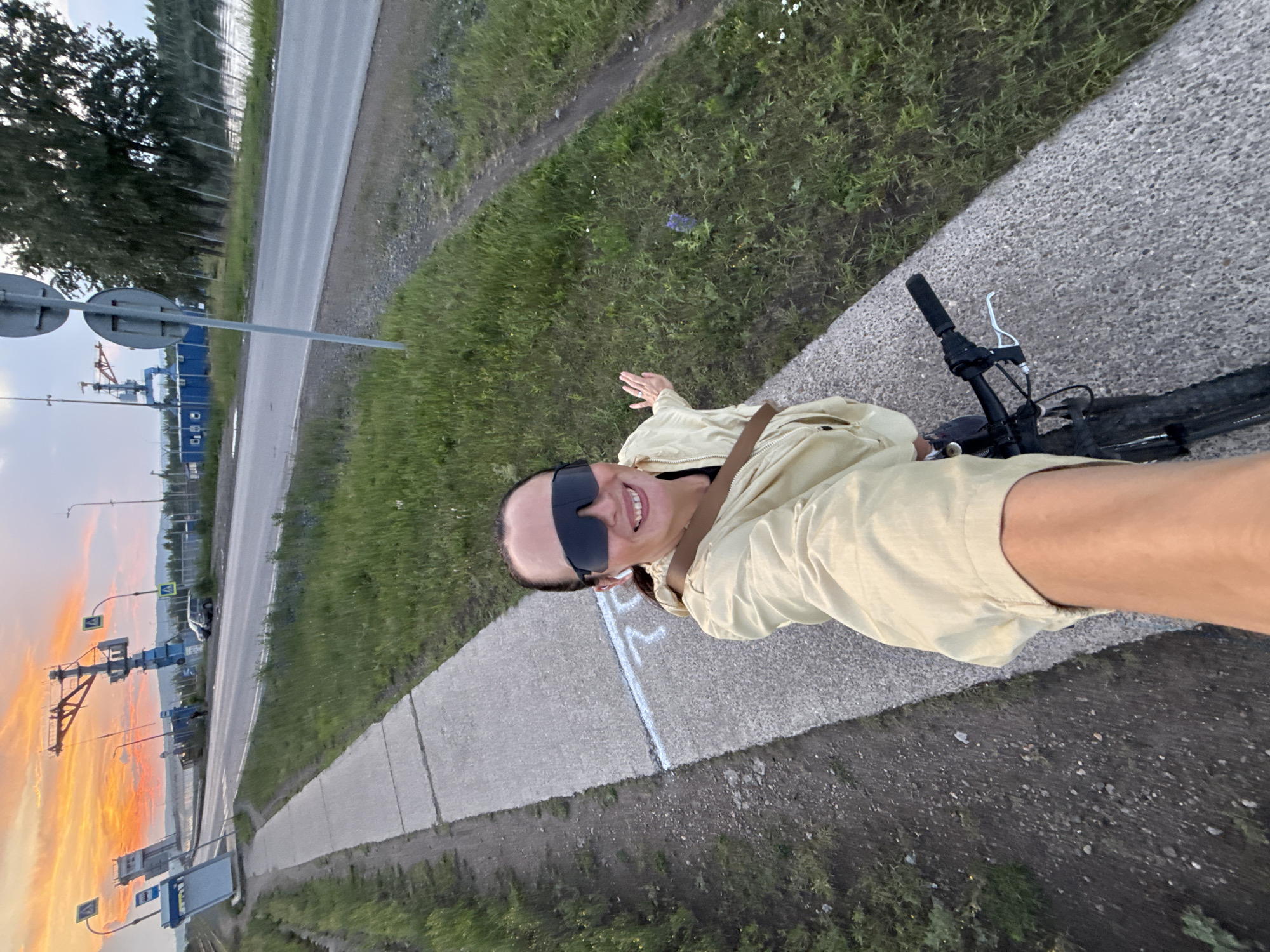 In Chaikovsky, Russia, on a paved path next to a road and grassy embankment, a woman is taking a selfie while riding a bicycle. She is wearing a tan jacket and large sunglasses, and she is smiling. The background shows a dramatic sunset with vibrant orange and pink clouds, a body of water, a blue industrial crane, and trees. There are also road signs and a vehicle visible.