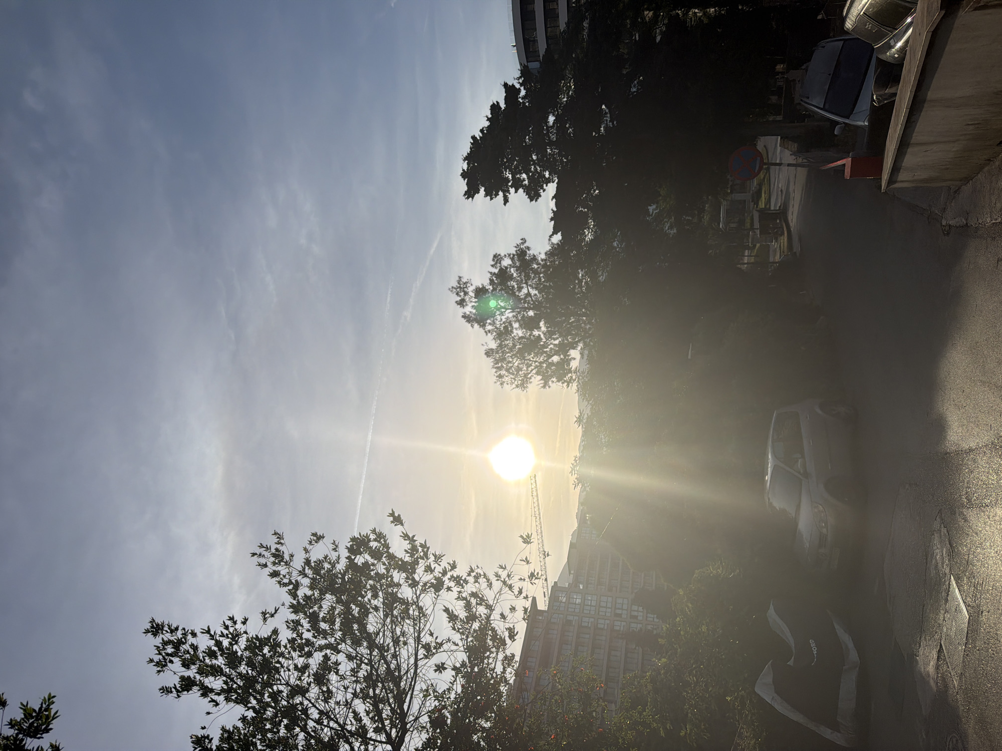 The image, taken in the city of Budva, Montenegro, depicts a street scene under bright sunlight. The sun is centrally located in the frame, obscured by haze, casting rays downward. In the foreground and midground, silhouetted trees frame the scene. The right side of the frame shows a paved road reflecting sunlight. A silver car is parked along the road. Further back, partially obscured by the glare of the sun, is a building with many windows, likely an apartment complex. A construction crane stands beside it. A 'No Stopping' sign is visible on the right side of the frame. The sky above has wispy clouds and a contrail.