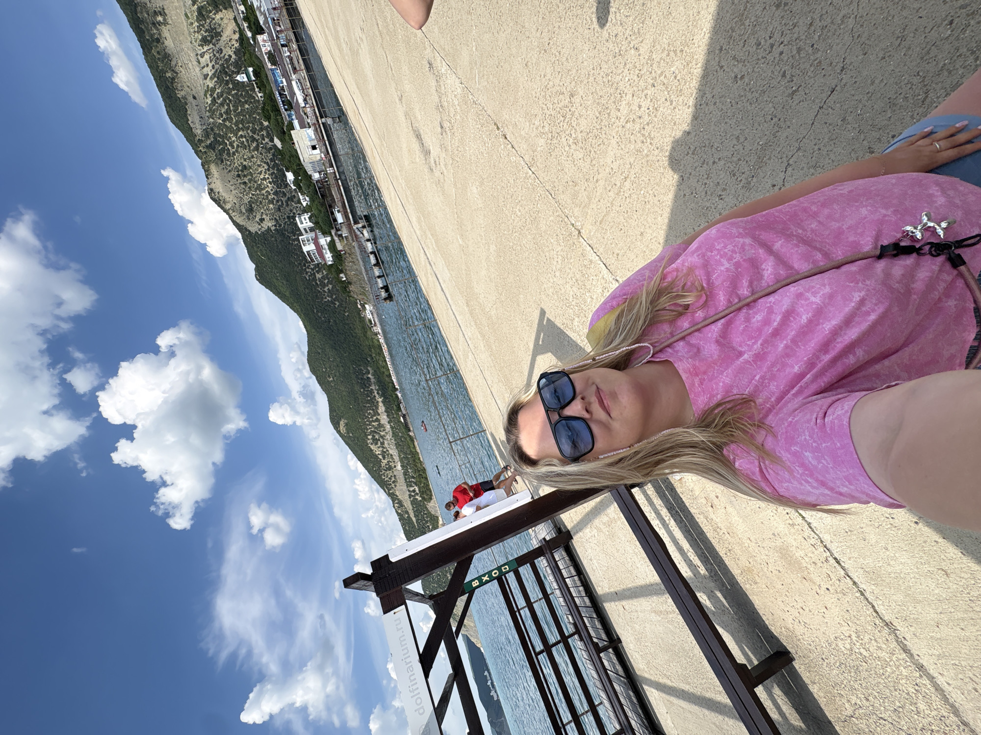 The image depicts a sunny coastal scene in Bol'shoi Utrish, Russia, featuring a woman taking a selfie on a concrete pier or promenade. The woman, positioned in the foreground, has blonde hair, wears blue rectangular sunglasses, and a pink, tie-dye pattern t-shirt. A light-colored beaded strap, possibly for her phone or glasses, is visible on her left side, and a silver balloon dog charm hangs from a dark strap across her chest.
Behind the woman, to the left, a brown wooden structure, likely an entrance or shelter for a pier, is visible. A white sign on this structure displays "dolfinarium.ru" and a vertically oriented green sign reads "ВХОД" (VKHOD, meaning "ENTRANCE") with an arrow pointing downwards. Beyond the structure and promenade is a calm blue body of water, likely the Black Sea, with a metal railing separating it from the walking area. Several small boats or kayaks are scattered on the water.
In the mid-ground, further down the promenade, two men are visible near the railing; one is wearing a red t-shirt. The background is dominated by large, green, arid-looking mountains that rise directly from the coastline. Numerous white and light-colored buildings, possibly hotels or resorts, are situated at the base of these mountains, along the water's edge. A small white structure with a green dome is visible higher on one of the mountain slopes. The sky above is clear blue with scattered, fluffy white cumulus clouds. Bright sunlight casts defined shadows on the concrete promenade.