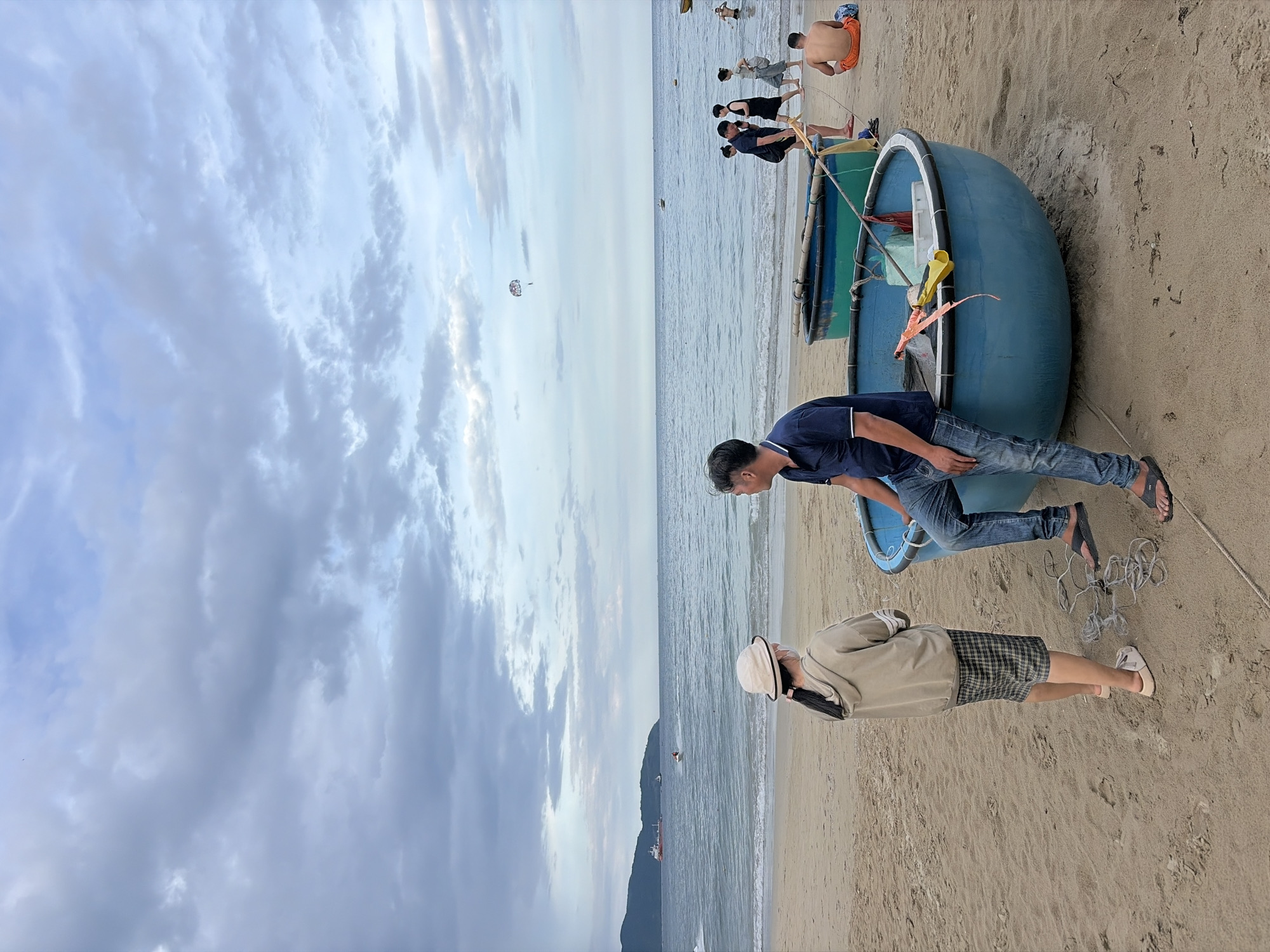 The image captures a beachfront scene at dusk in Đà Nẵng, Vietnam. In the foreground, a man in a dark blue polo shirt and jeans sits on the edge of a round, blue basket boat, facing away from the viewer and towards the ocean. He is wearing black flip-flops. To his left, a young woman wearing a light brown hoodie, plaid shorts, and a white hat with a mask stands with her back to the camera, also looking out at the sea. She is wearing white sandals.
The sand is a light brown, and several ropes are coiled on the ground near the boats. Another basket boat, painted green and blue, is partially visible behind the blue one.
In the middle ground, the calm ocean stretches to the horizon under a cloudy sky. Waves gently lap the shore. Several people are visible on the beach and in the water. A few individuals are walking along the shoreline, and one person is seated on the sand wearing orange shorts. Further out in the water, a jet ski is being ridden, and a parasail is visible in the sky. A large red and white ship is docked in the distance, near a mountainous coastline. The overall atmosphere suggests a peaceful, end-of-day scene at a coastal resort.