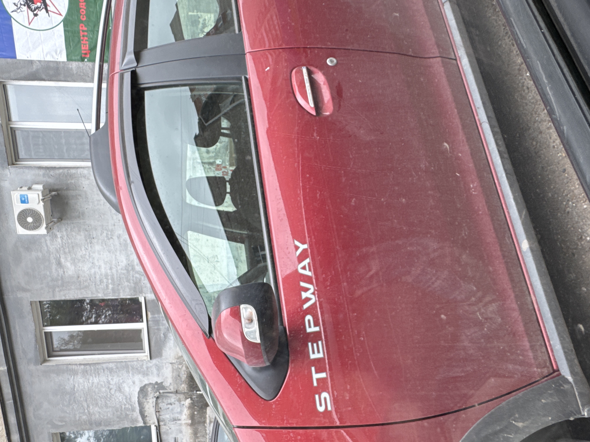 A red car, with the word "STEPWAY" in white lettering on its side, is parked on a street in Ufa, Russia. The car is dirty, with dust and scratches visible on its surface. A black plastic trim runs along the bottom of the car. In the background, a building with a grey textured facade and a window is visible. An air conditioner is mounted on the wall of the building. A banner with red and white lettering is also present in the background.