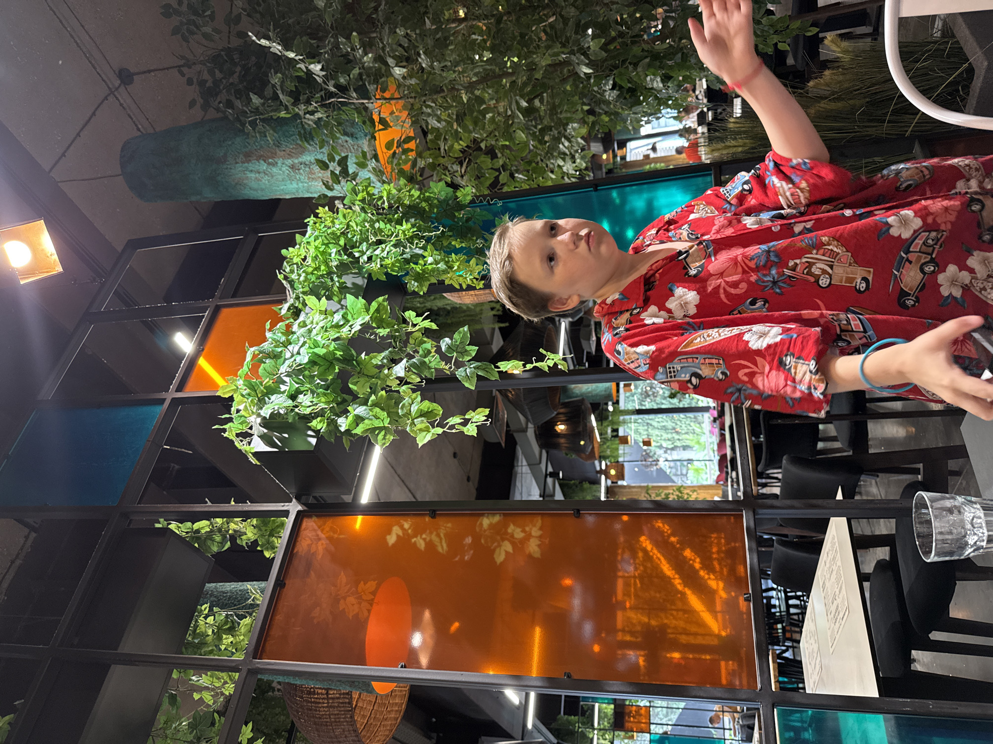 A boy with short brown hair is sitting at a table in a restaurant. He is wearing a red shirt with a Hawaiian print that features cars and flowers. The boy is looking off to the right and has his right arm extended. He is wearing a blue bracelet on his left wrist. In the foreground, a glass of water is on the table. The restaurant has a modern design with a black metal frame separating colored glass panels. There are hanging plants and tall, tree-like decorations. In the background, other tables and patrons are visible. The location is identified as a restaurant in Yalta, Ukraine.