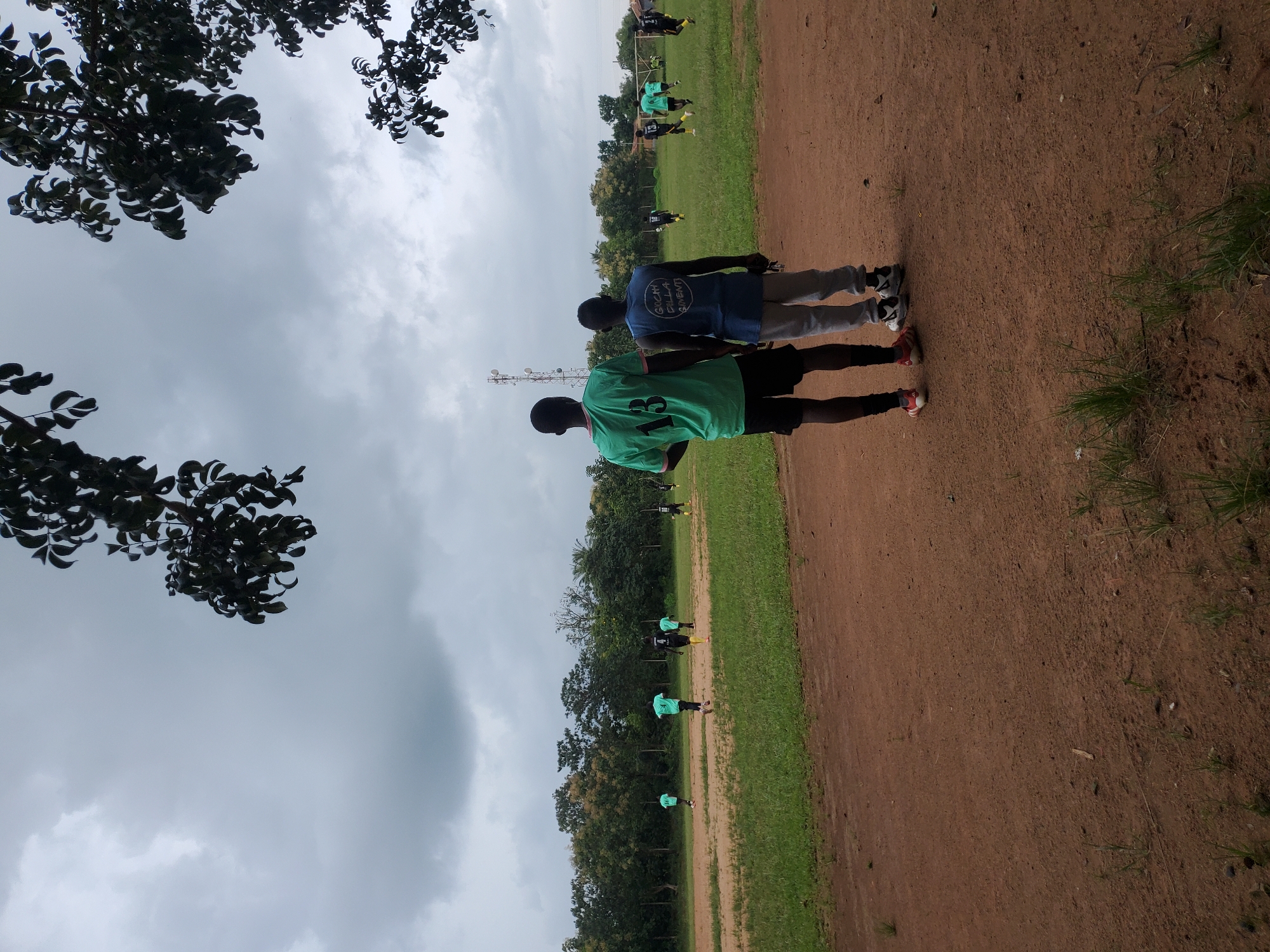 Two young Black men are standing on a dirt field, facing away from the camera. The man on the left is wearing a green t-shirt with the number "13" on the back and black shorts. The man on the right is wearing a blue sleeveless shirt with writing on the back and light gray sweatpants. They are looking towards a grassy field where a soccer game is in progress. Several other young Black men, dressed in athletic wear, are visible on the field, some playing soccer and others observing. The field is surrounded by trees and vegetation, and the sky is overcast. The location is identified as a soccer field in Miango, Nigeria.