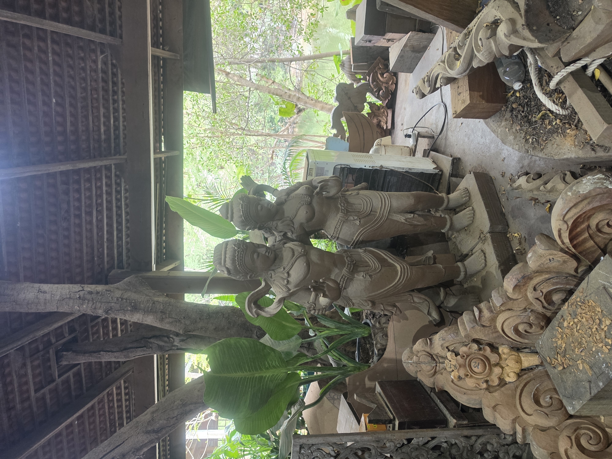 This media file captures a semi-outdoor artisan's workshop or display area in Pattaya City, Thailand, illuminated by natural daylight.
At the core of the scene are two ornately carved statues, likely made of stone or concrete, depicting figures resembling deities or traditional characters. They stand side-by-side, adorned with detailed headdresses, elaborate garments, and distinct hand gestures; one appears to hold a staff or serpent-like object. Lush green tropical plants are integrated into the display around their bases.
The area is sheltered by a rustic structure featuring large wooden beams and a corrugated tile roof visible from below. A prominent, gnarled tree trunk is incorporated into the left side of the architectural framework.
To the right of the statues, a concrete floor is cluttered with various wooden carvings, some appearing unfinished. These include large, scrolled decorative pieces and blocks of wood. Wood shavings on one carving suggest ongoing work. Amidst these items, a clear plastic bottle, a coil of rope, and parts of an outdoor air conditioning unit are visible. A specific circular carved wooden element displays the number "21" inscribed within it.
Beyond the sheltered workspace, a vibrant natural environment unfolds. Dense green trees and foliage dominate the background, with glimpses of a body of water, possibly a river or canal, visible through the leaves. The overall atmosphere suggests a humid, tropical climate and a dedicated space for traditional craftsmanship.