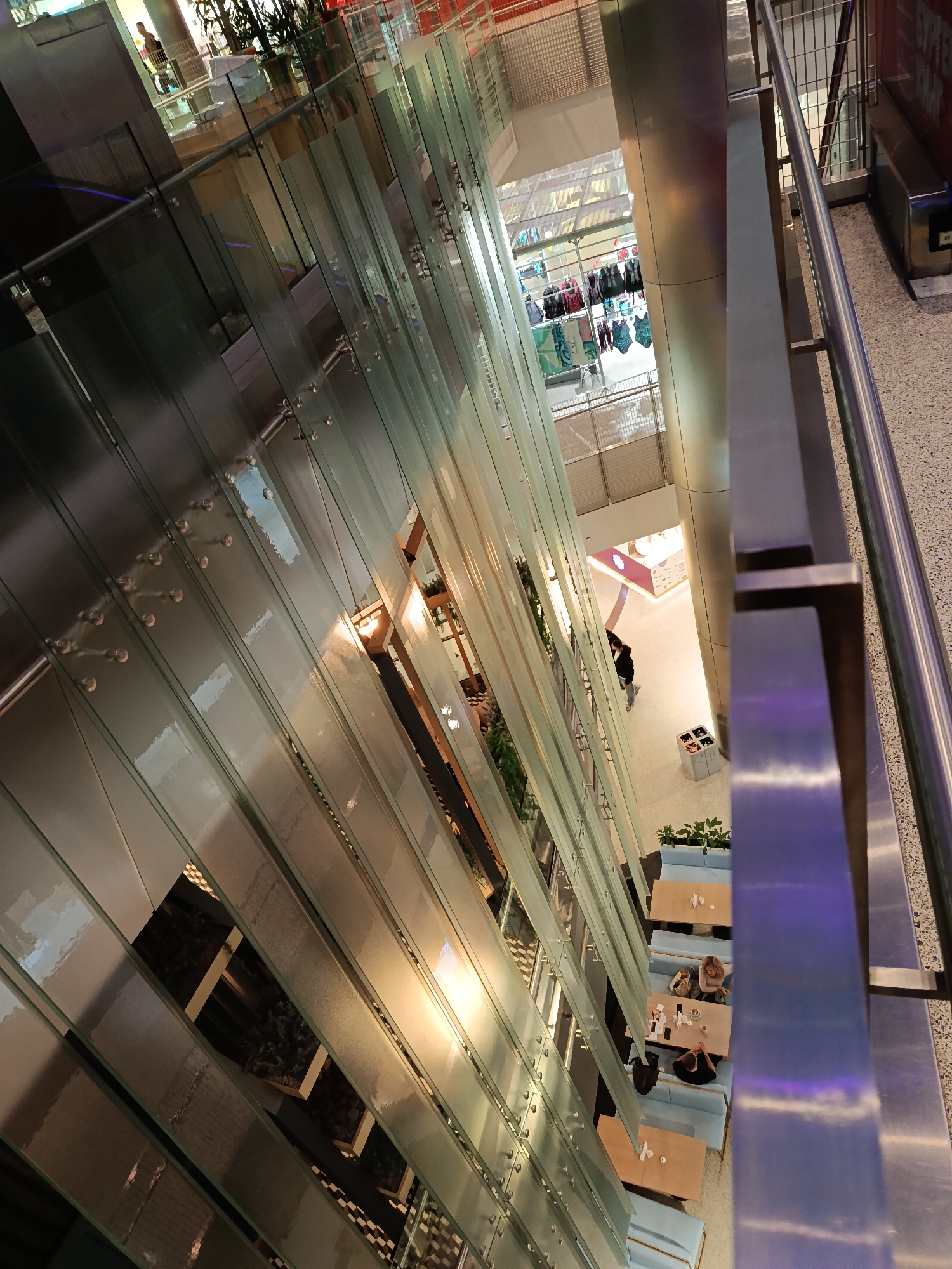 The image depicts an interior view from a high vantage point, possibly overlooking several floors of a shopping center or atrium. The scene is dominated by a structure of glass panels or partitions, aligned vertically and secured with metallic fixtures. These panels create a visual barrier or separation while allowing light and views to pass through.
Below, there is a restaurant or cafe area with tables and seating arrangements. People are visible at some of the tables, appearing to be engaged in dining or conversation. Further away, there are retail displays, including what looks like a swimsuit or beachwear section, indicating a shopping environment. A lone individual walks through the lower level.
The viewpoint is from an upper level with a metal railing. The lighting appears to be a combination of natural and artificial sources, with a warm glow illuminating some areas and a cooler tone in others. The overall atmosphere is modern and airy, suggestive of a contemporary commercial space. While the city of Moscow, Russia cannot be determined from image data only, this cannot be confirmed.
