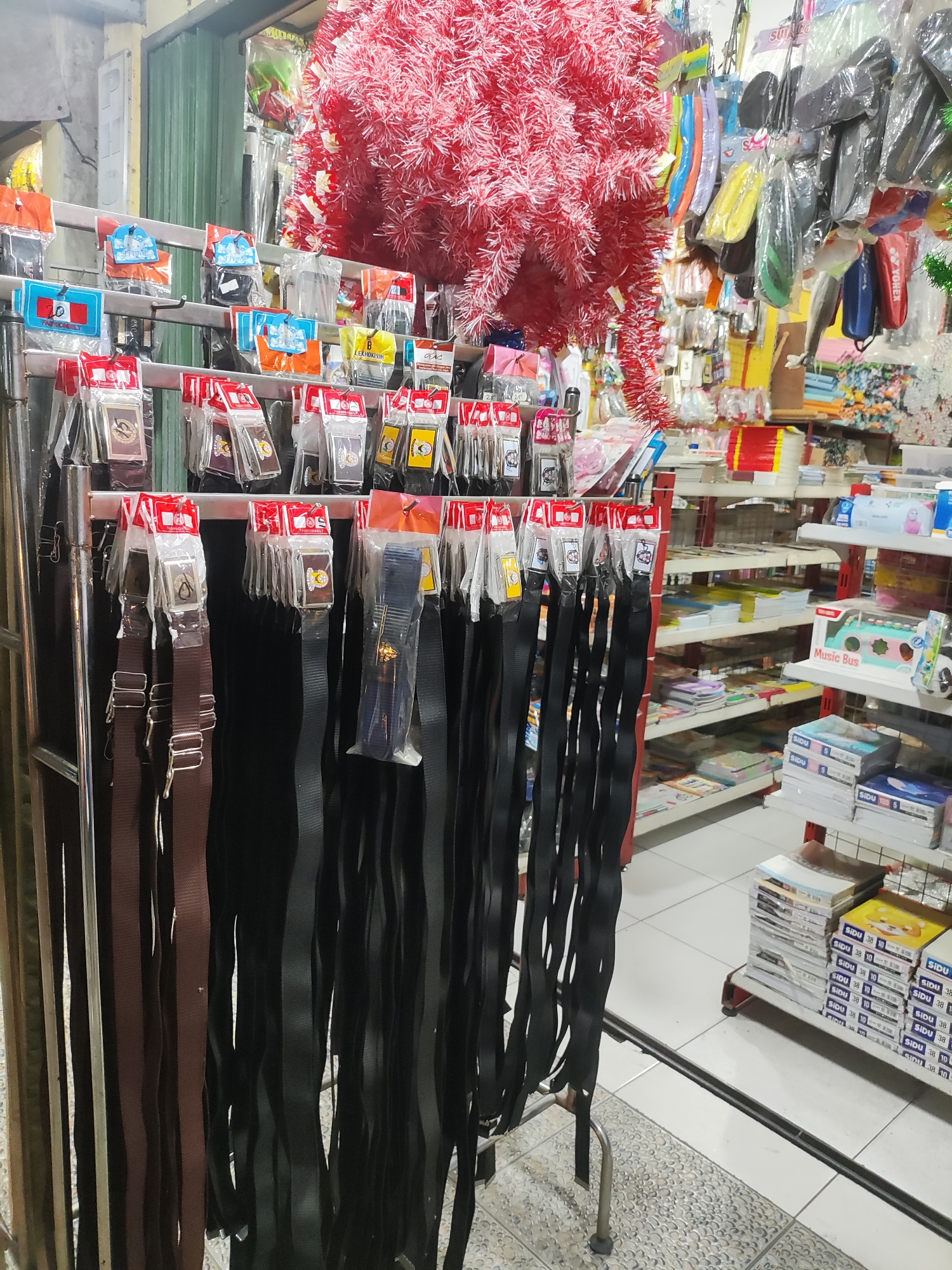The image shows a retail store in Wanajaya, Indonesia, likely a small shop or a stall within a larger market. The foreground is dominated by a display of belts hanging from a metal rack. These belts appear to be of various colors, including brown and black, and are packaged with cardboard headers. Some of the belts have decorative buckles. Behind the belts, various other items are visible, including what appears to be small decorative pins or keychains packaged in plastic, and a large cluster of red tinsel or garland hanging from the ceiling, suggesting a festive or seasonal display. Further in the background, shelves are stocked with various goods, including what look like school supplies, books, and possibly stationery items. The overall impression is of a bustling local shop selling a diverse range of affordable merchandise. The floor appears to be tiled.