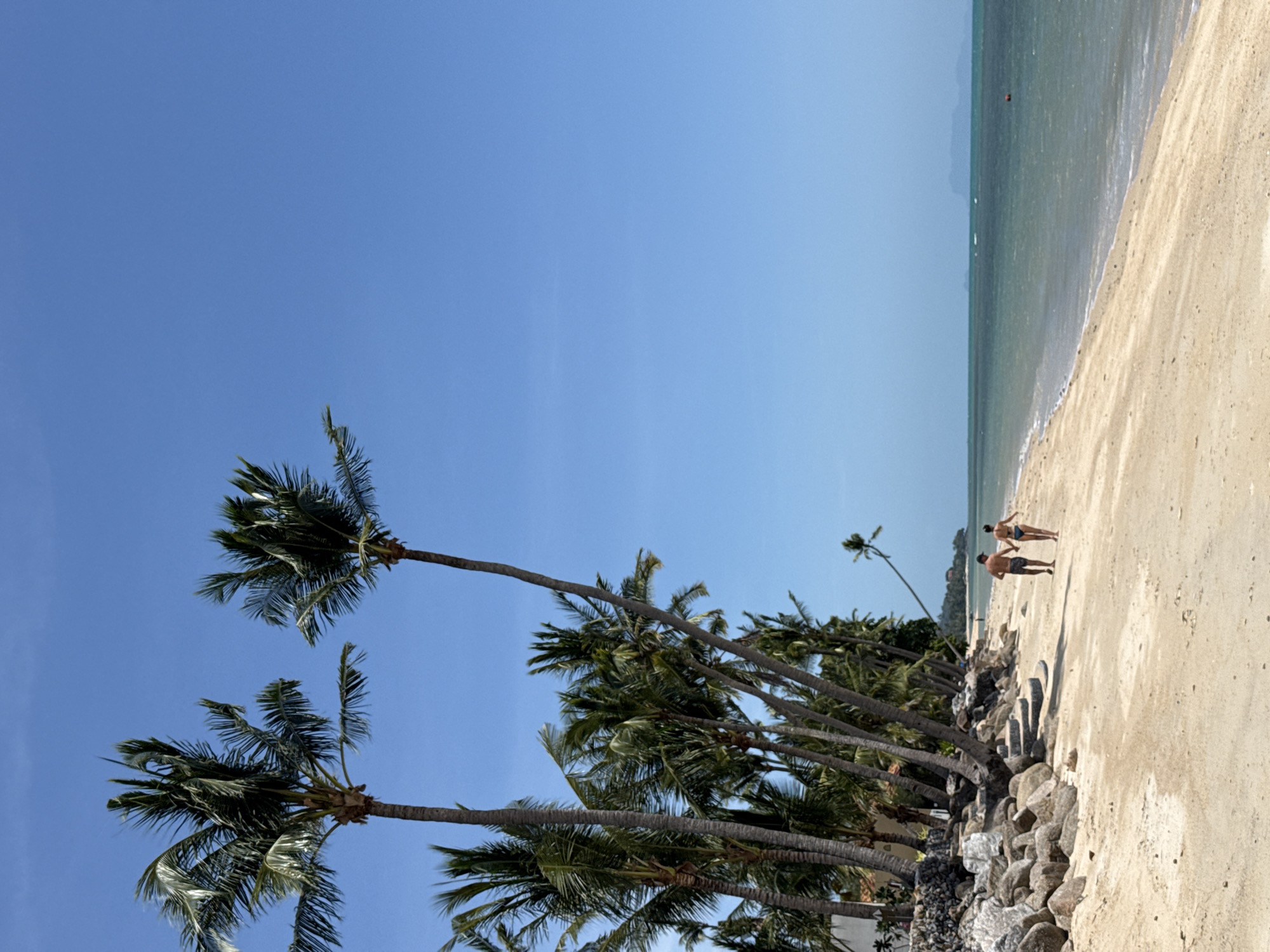 A couple walks hand-in-hand on a sandy beach in Ko Samui District, Thailand. The man is wearing dark swim trunks, and the woman is wearing a bikini. They are walking away from the camera towards the water. The beach is lined with palm trees, and there are rocks and a stone staircase leading up from the beach on the left side of the image. The ocean is calm and blue, with a distant island visible on the horizon. The sky is clear and blue.