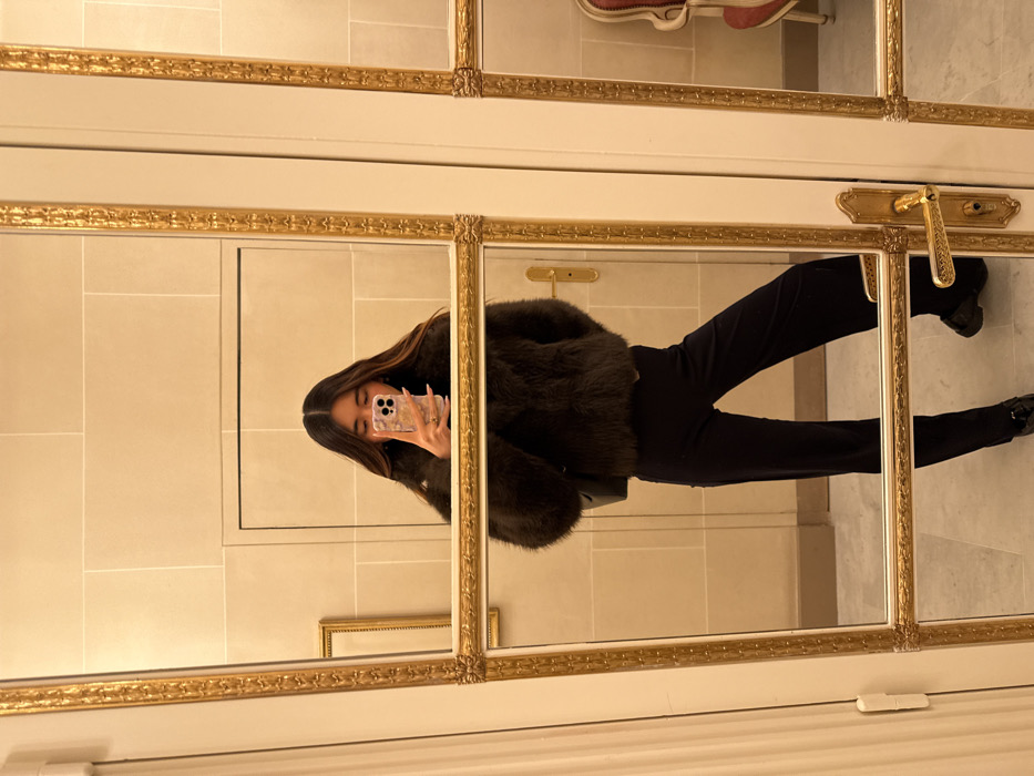 The image captures a full-length mirror selfie of a person in what appears to be an elegant interior. The original image is rotated 90 degrees clockwise, so the person appears to be lying on their back, but in an upright orientation, they would be standing.
The person, whose face is largely obscured by the smartphone they are holding, has long, straight, dark brown hair that falls over their shoulders. They are wearing a dark, possibly black or very dark brown, fluffy or faux fur jacket, which has a substantial volume. Below the jacket, they are wearing dark, form-fitting trousers or leggings. Their feet are clad in dark, possibly black, shoes. The hand holding the phone has manicured nails painted in a light color, possibly with delicate patterns or embellishments. The smartphone has a light-colored, possibly white or cream, case with a subtle floral or decorative design.
The setting is characterized by a large, multi-paneled mirror that serves as the backdrop for the selfie. The mirror panels are divided by thick, ornate gold frames, featuring intricate carvings and decorative elements at the corners and along the edges. The reflection in the mirror shows a light-colored wall, possibly made of large, pale stone tiles or panels, with visible grout lines or seams. In the upper right corner of the reflected background, an ornate golden door handle or fixture is visible. Further down and to the left in the reflection, a smaller, empty golden picture frame is seen hanging on the wall. The overall ambiance suggested by the mirror's design and the reflected background is one of classic elegance, potentially in a hotel, a stately home, or a luxurious public space.
The primary activity is the person taking a self-portrait using their smartphone, reflected in the ornate mirror. There is no visible text in the image.