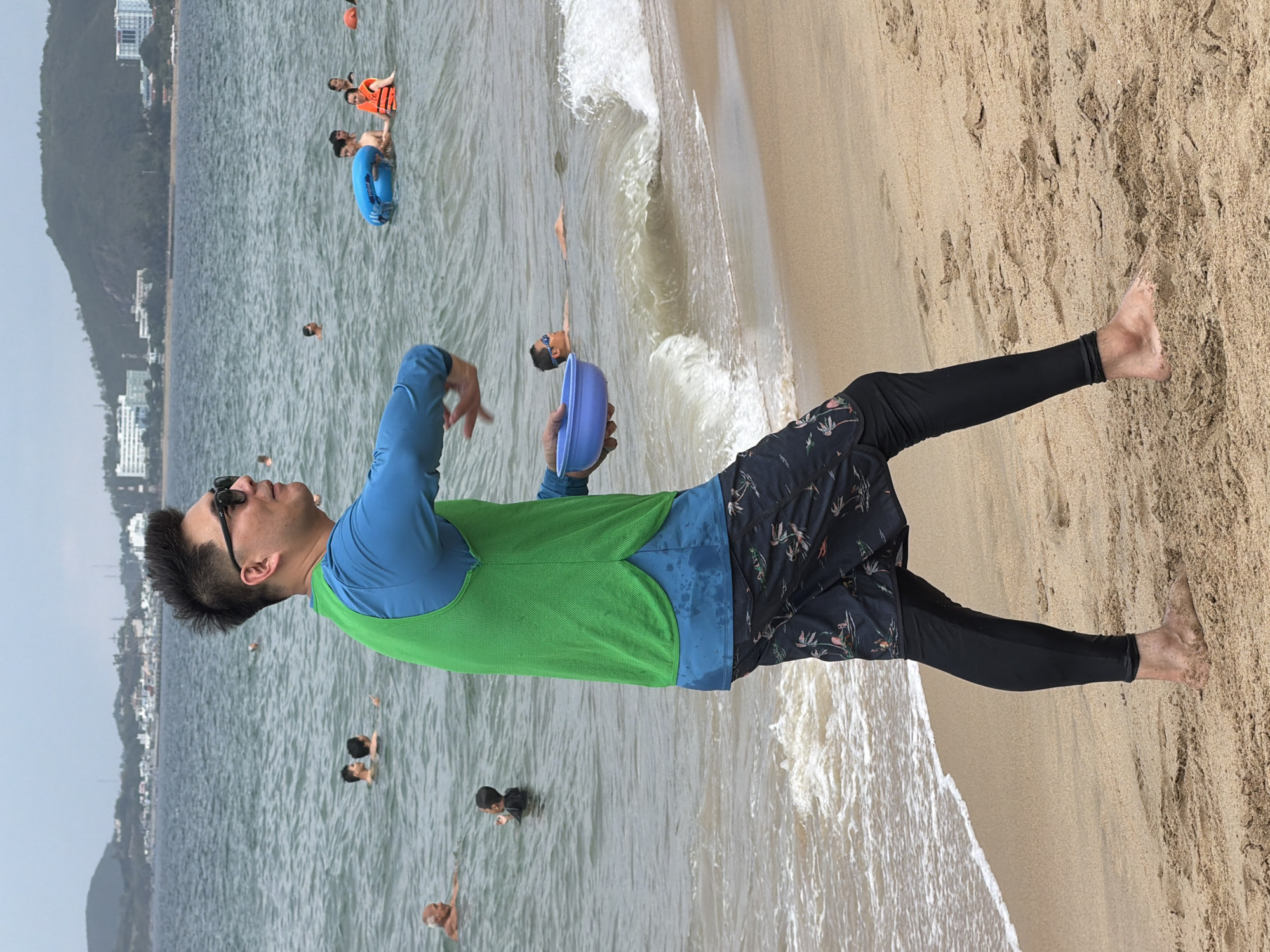 A man in a blue long-sleeved shirt and a green vest stands on a sandy beach in Nha Trang, Vietnam, holding a blue bowl. He is wearing dark swim shorts over black leggings and sunglasses, with his hair spiked up. His right arm is extended, and he appears to be in the process of throwing something from the bowl. In the background, the ocean is visible with several people swimming and playing in the water, some wearing life vests. The shoreline features gentle waves lapping onto the sand. In the distance, a hilly coastline with buildings and trees can be seen under an overcast sky. The overall scene captures a moment of activity and recreation at the beach.