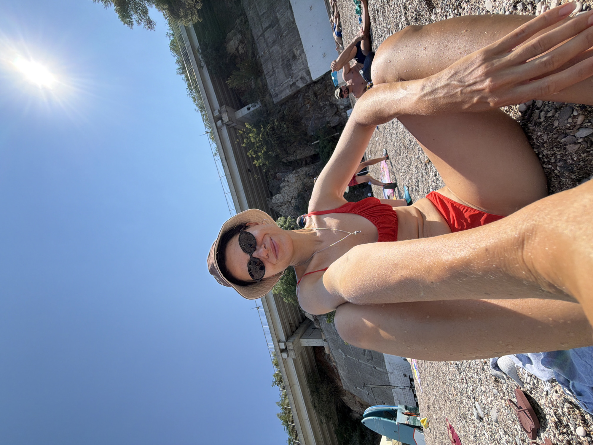 The image shows a woman in a red bikini and a bucket hat sitting on a pebble beach, facing the camera. She is wearing sunglasses and a silver necklace with a cross pendant. Her legs are spread apart and her hands are resting on her thighs. The sun is shining brightly in the sky, casting lens flares. In the background, there is a large concrete structure with a bridge on top, covered in greenery. Several other people are visible on the beach, some lying down and others walking. There are also two paddleboards leaning against a fence on the left side of the image. The overall scene suggests a sunny day at a beach with some man-made infrastructure in the background.