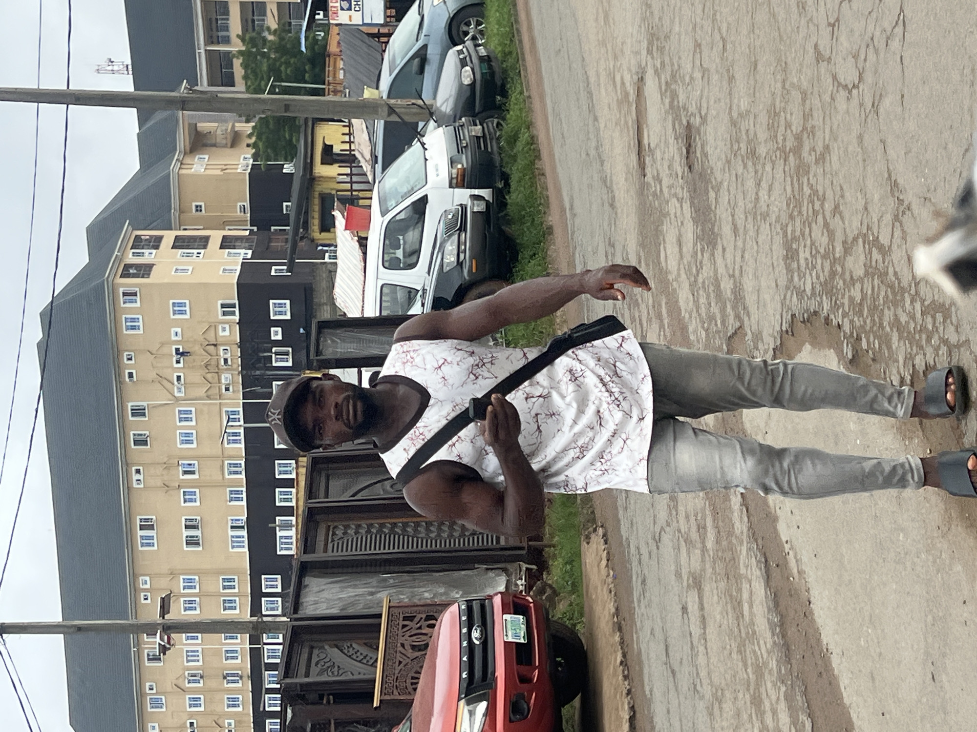 In Nkpor, Nigeria, a Black man walks on a cracked asphalt road, holding a smartphone. He wears a white tank top with a red marble-like pattern, gray denim jeans, and black sandals. A black crossbody bag is slung over his right shoulder. Behind him, parked vehicles, including a red Ford Ranger pickup truck, a black sedan, a white van, and a blue sedan, line a grassy median. In the background, a large, tan, multi-story building with dark roof tiles stands prominently, featuring numerous windows with blue frames. The sky is overcast.