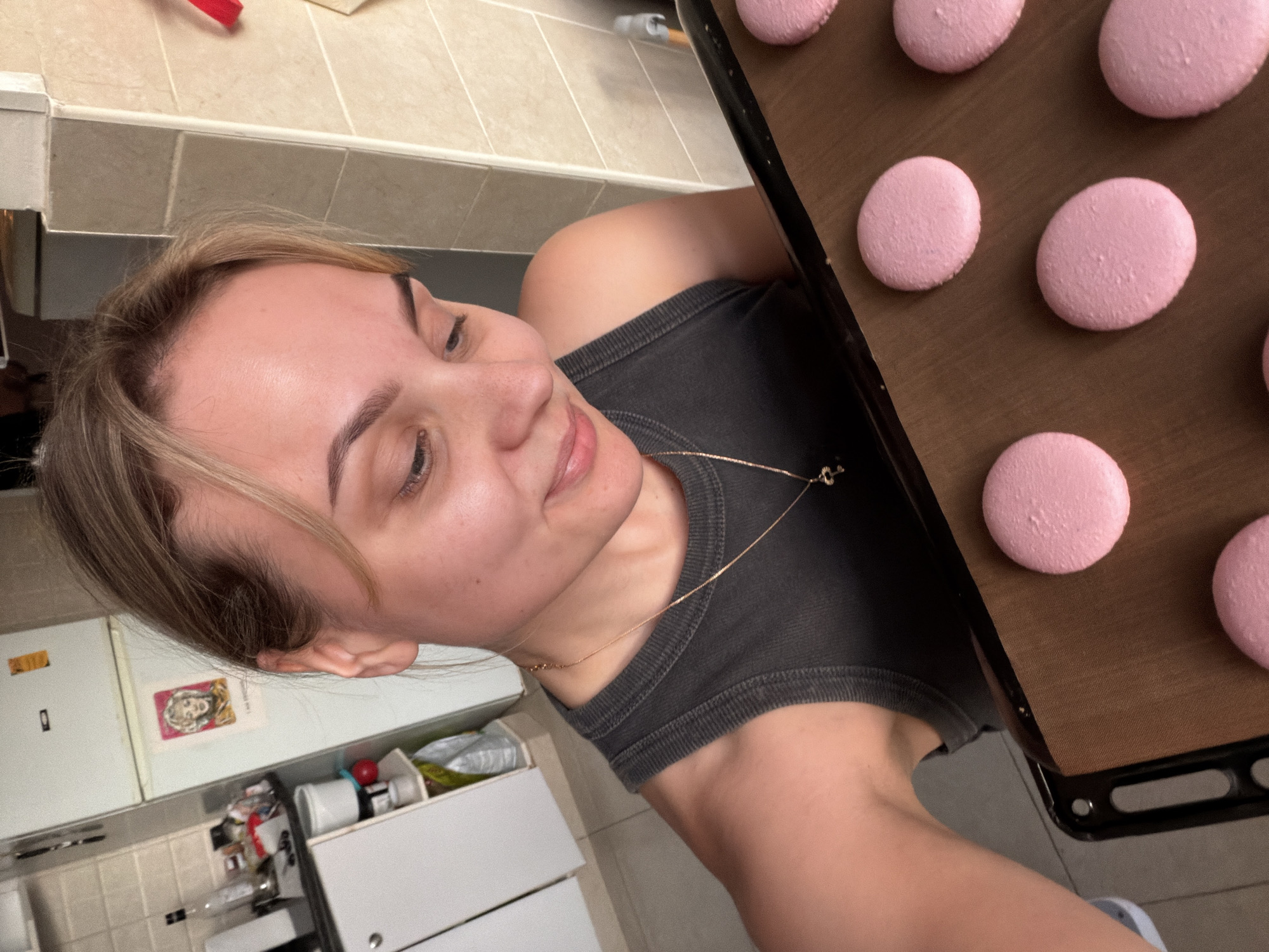 This rotated eye-level shot captures a young woman in a domestic kitchen in Tel-Aviv, Israel, engaged in baking. The woman, with light brown hair and a pleasant, slightly smiling expression, is positioned on the left side of the frame, looking towards a baking sheet filled with pink macarons. She wears a dark grey ribbed tank top and a thin golden chain necklace.
The activity clearly visible is the preparation of macarons, with several perfectly piped, unbaked pink macarons neatly arranged on a brown silicone baking mat on a black baking tray, positioned on the right side of the image.
The setting is a home kitchen, characterized by light-colored tiled walls and countertops, and white kitchen cabinets or drawers visible in the background on the lower left. Above these cabinets, a white refrigerator is partially seen, adorned with a small picture or drawing of a stylized face and some barely legible text next to it. The kitchen appears lived-in, with various bottles, containers, and other items loosely arranged on the counter and inside partially open drawers, suggesting an ongoing cooking or baking session. The woman's expression conveys a sense of contentment or focus on her baking task. The time of day is not discernible, as the scene is lit by indoor lighting.