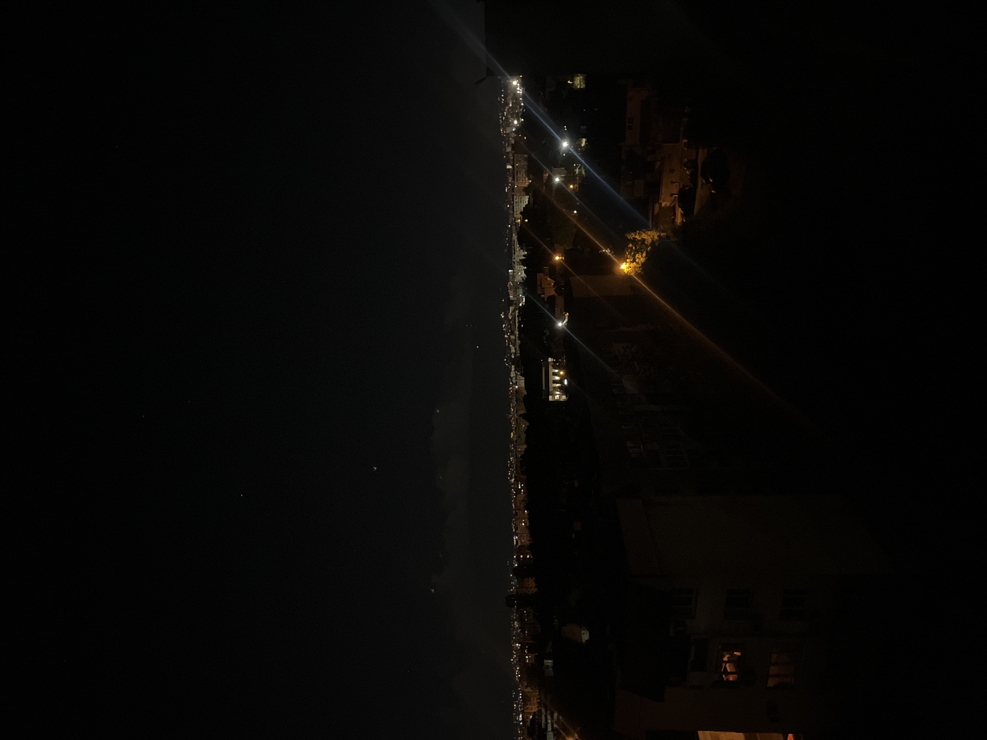 The image shows a nighttime view of the coastline. In the right portion of the image are buildings and houses, with streetlights illuminating the streets, and windows visible. The left portion is a body of water, such as the sea or ocean. It is dark, and the light from the coast reflects off the water. The location is not identifiable as Sofia, Bulgaria from the image alone.