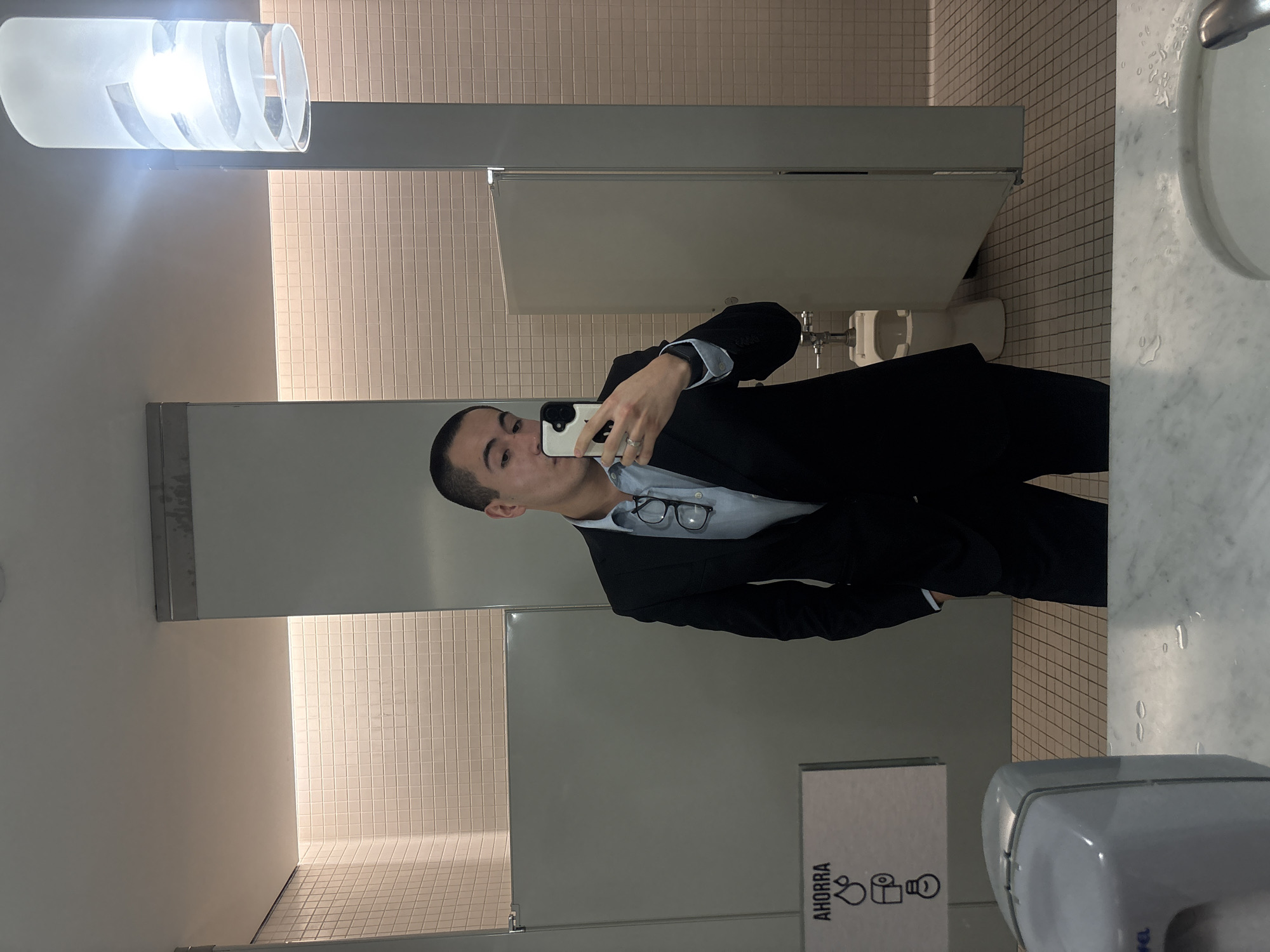 A young person, dressed in a dark suit jacket over a light blue collared shirt, takes a mirror selfie in what appears to be a public restroom. The individual has short, dark hair and is holding a white smartphone, partially obscuring their face, while making a slight pucker with their lips. A pair of glasses is tucked into the front of their shirt. A ring is visible on their right ring finger.
The setting is brightly lit by artificial light, including a modern, cylindrical light fixture with a translucent, swirled design mounted above the mirror on the left. The restroom features light-colored, small square tiles on the walls and floor, creating a grid pattern. Behind the person, several light gray toilet stall doors are visible, with a white toilet faintly seen inside one.
To the right, a white marble countertop with a sink basin and a metal faucet is present, with water droplets visible on the surface, suggesting recent use. Below the mirror, a gray plastic hand dryer is partially visible, next to a white sign with black text and icons. The sign prominently displays the Spanish word "AHORRA" (meaning "SAVE"), followed by icons depicting a dripping faucet (representing water conservation) and a toilet paper roll or tissue box, and a lightbulb, indicating a general conservation message. The overall impression is a clean, well-maintained commercial or institutional restroom.