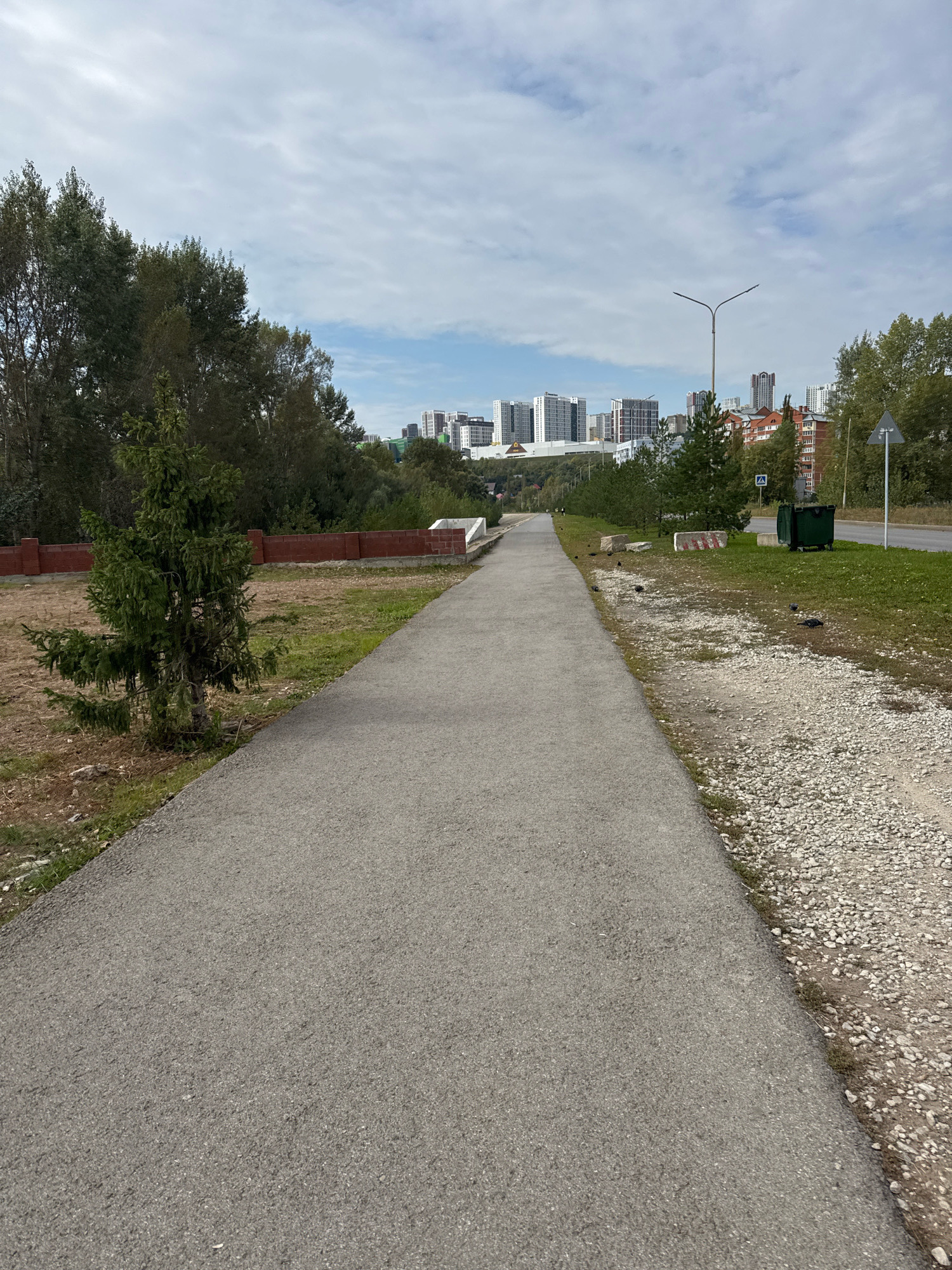 The image depicts a paved pathway, likely a pedestrian or bicycle lane, curving gently to the right and extending into the distance. The pathway is bordered by a strip of gravel and then grass on the right, and by a grassy area with a young spruce tree and a low brick wall on the left. In the background, a cluster of modern apartment buildings of varying heights and architectural styles rises against a cloudy sky. A street lamp is visible on the right side of the pathway, along with a green trash receptacle and a traffic sign. The overall scene suggests an urban park or a newly developed area on the outskirts of a city, consistent with the description of Ufa, Russia.