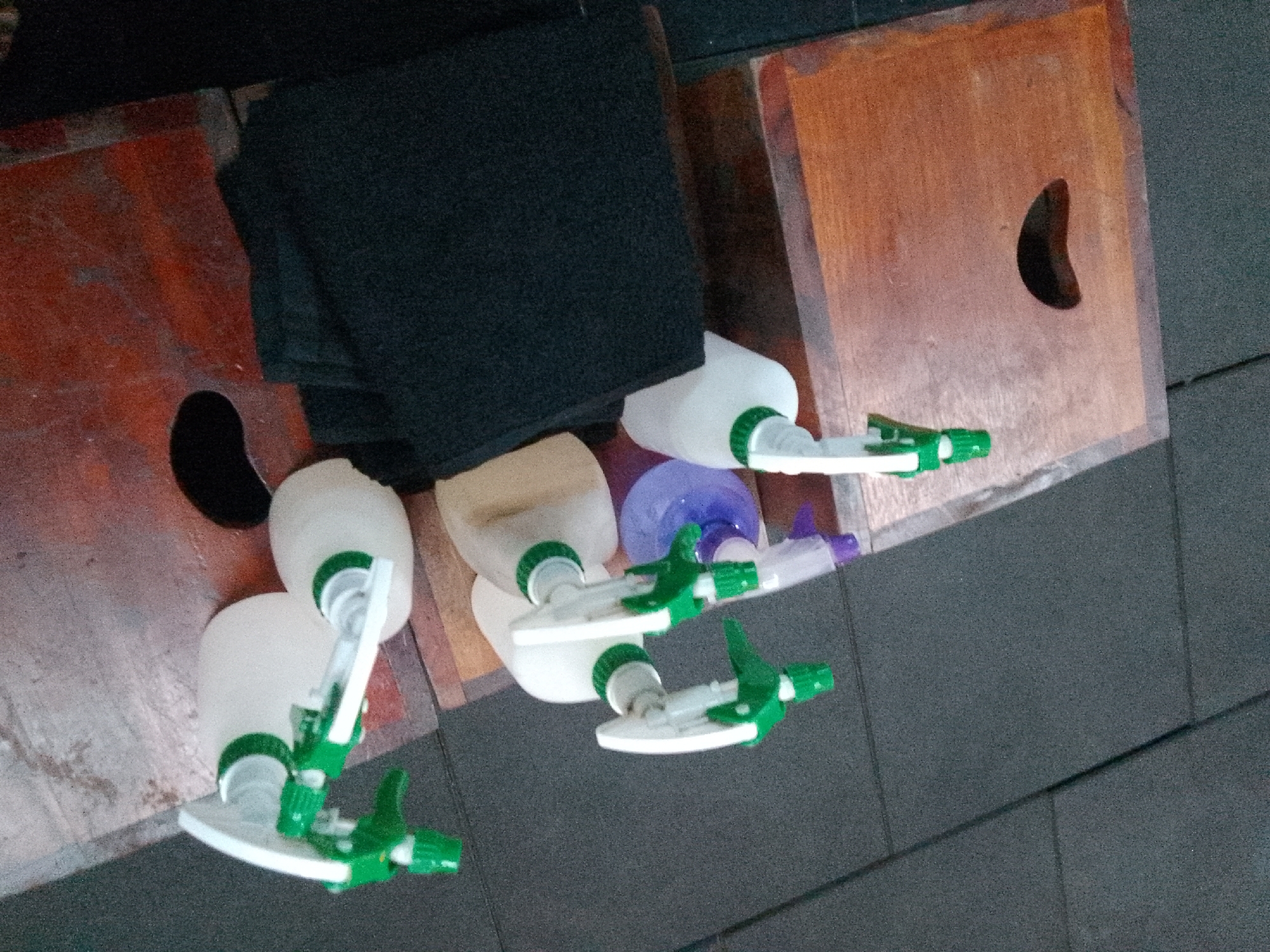 The image displays a collection of white plastic spray bottles with green trigger mechanisms arranged on a textured, dark gray surface. Some of the bottles are partially obscured by a folded dark fabric. The bottles are positioned on what appears to be wooden surfaces. One wooden surface is a reddish-brown color and has a dark, oval-shaped hole. Another wooden surface is a lighter, orangey-brown color and also has a dark, curved opening. The overall scene is indoors or under a sheltered area, given the lighting and the nature of the objects. There are no people visible in the image, and no specific actions or interactions are depicted. The context of Trenggalek, Indonesia, is not visually ascertainable from the provided image.