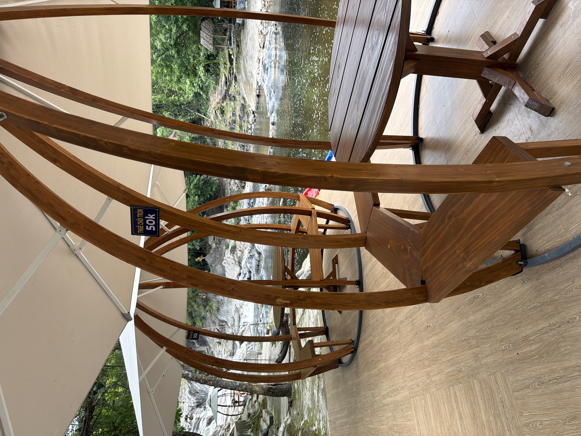 This image depicts an outdoor seating area located in Khanh Vinh, Vietnam, situated next to a river. The area features several wooden, pod-like structures with curved wooden frames and white canopies, providing shade. Each pod contains a wooden table and benches. In the foreground, a table and benches are clearly visible. In the background, the river flows over rocks, and a stone path leads up to a small hut with a thatched roof. Some people are visible in the distance, near the river and on the rocky terrain. A sign in Vietnamese, reading "THUÊ CHỖ TRÒN 50K", which translates to "RENT THE WHOLE PLACE 50K", is attached to one of the wooden frames. The ground is paved with wooden planks. Lush green trees and vegetation surround the scene, creating a natural and serene environment.