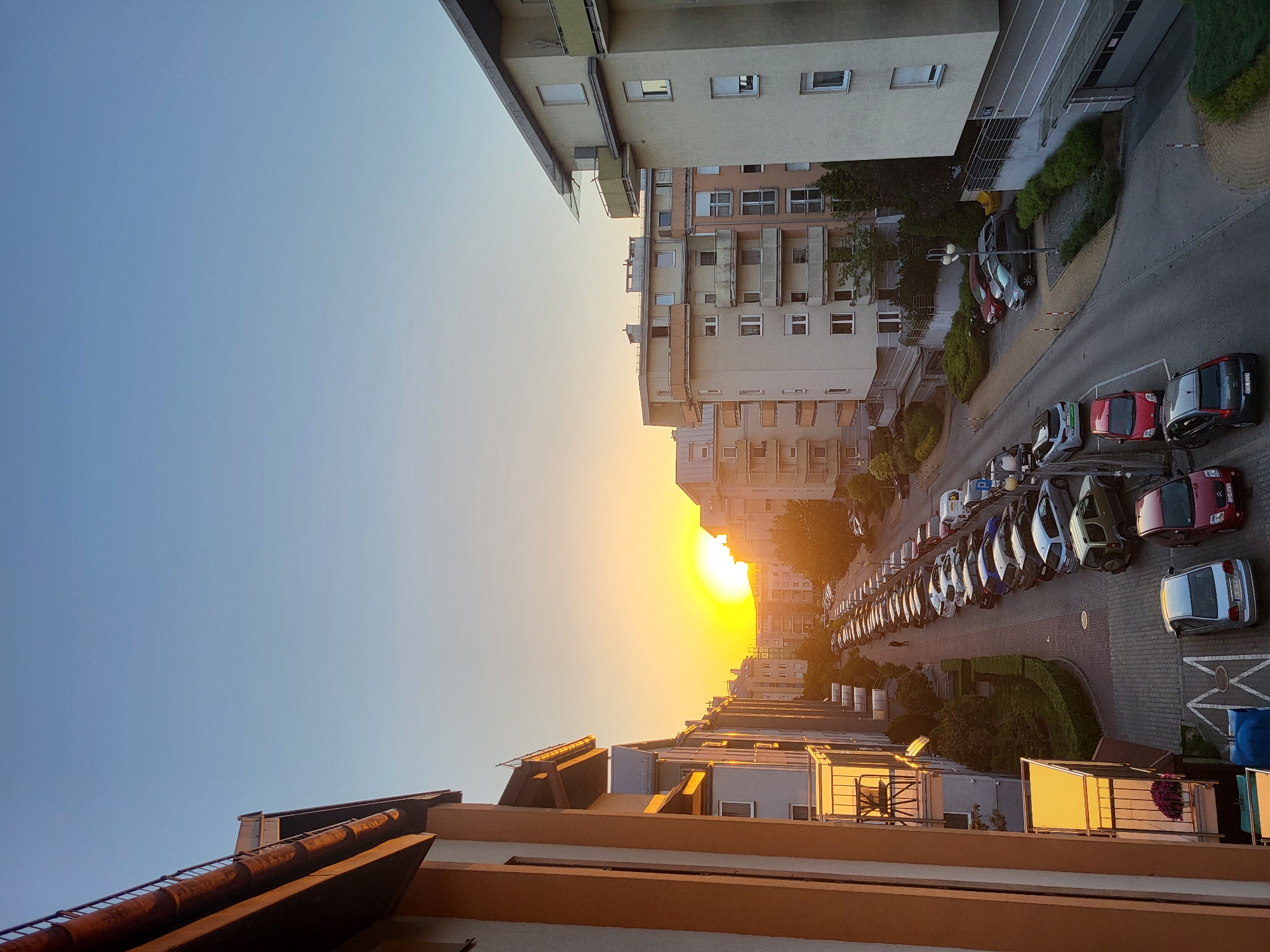 The image shows a view of an urban street at sunset. Tall buildings line both sides of the street, with rows of parked cars filling the parking spaces along the sides. The sun is setting in the distance, casting a warm golden glow over the scene. The sky is a gradient of blue and orange near the horizon. The location is Unknown, Unknown. There are no people visible.